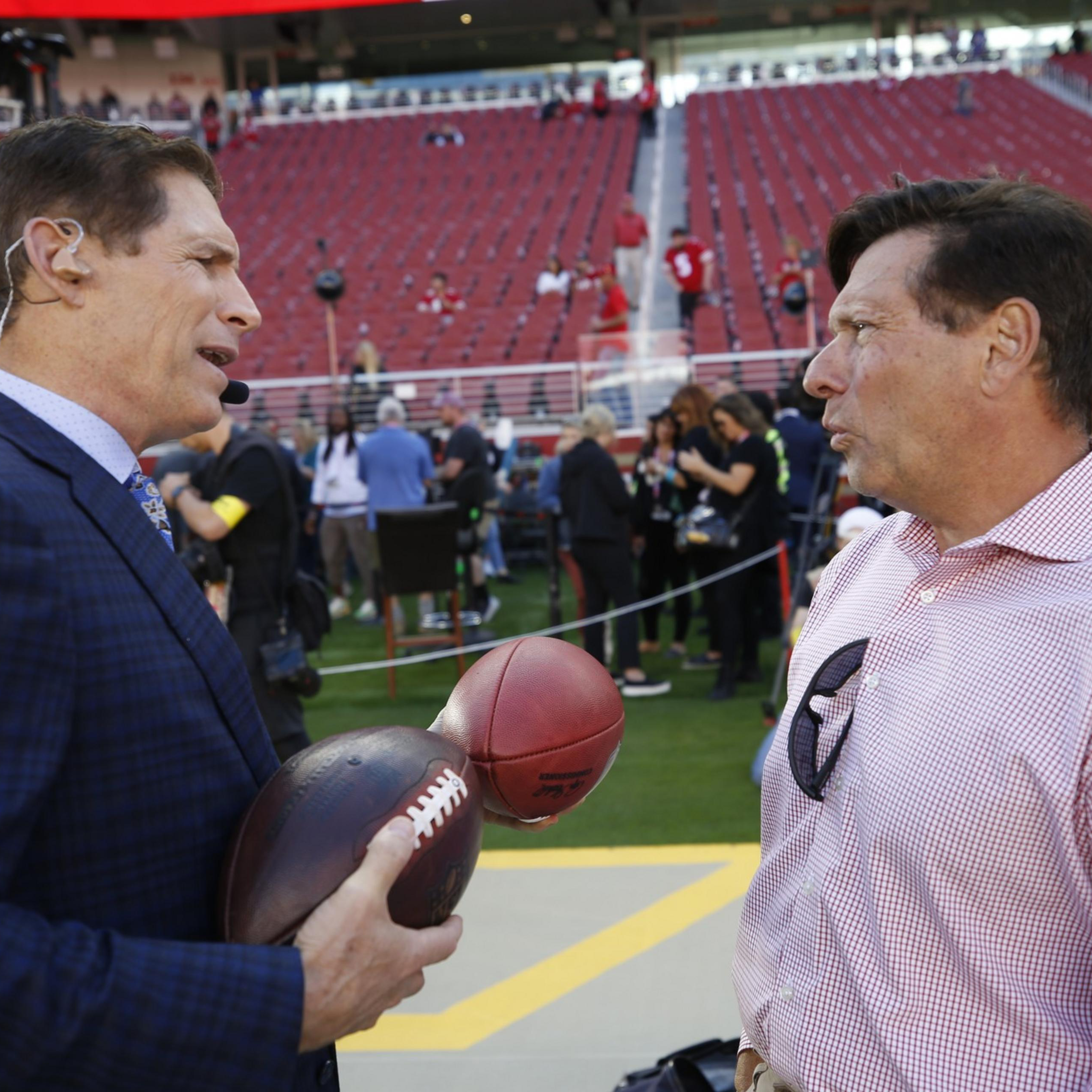 Two men talk on a football field near the stands, one wearing a suit with a headset holding two footballs, the other in a checkered shirt with glasses hanging from it.