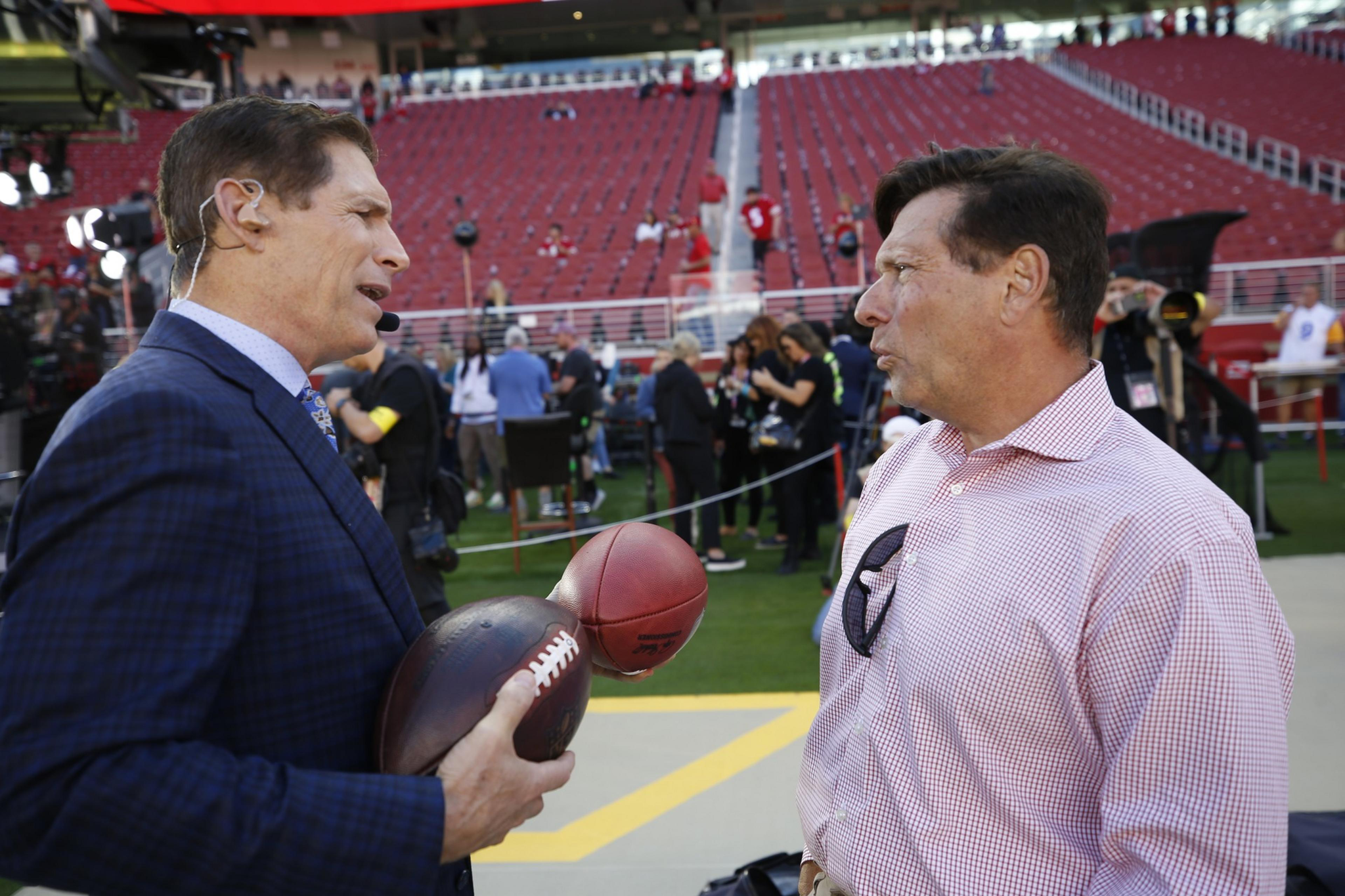Two men talk on a football field near the stands, one wearing a suit with a headset holding two footballs, the other in a checkered shirt with glasses hanging from it.