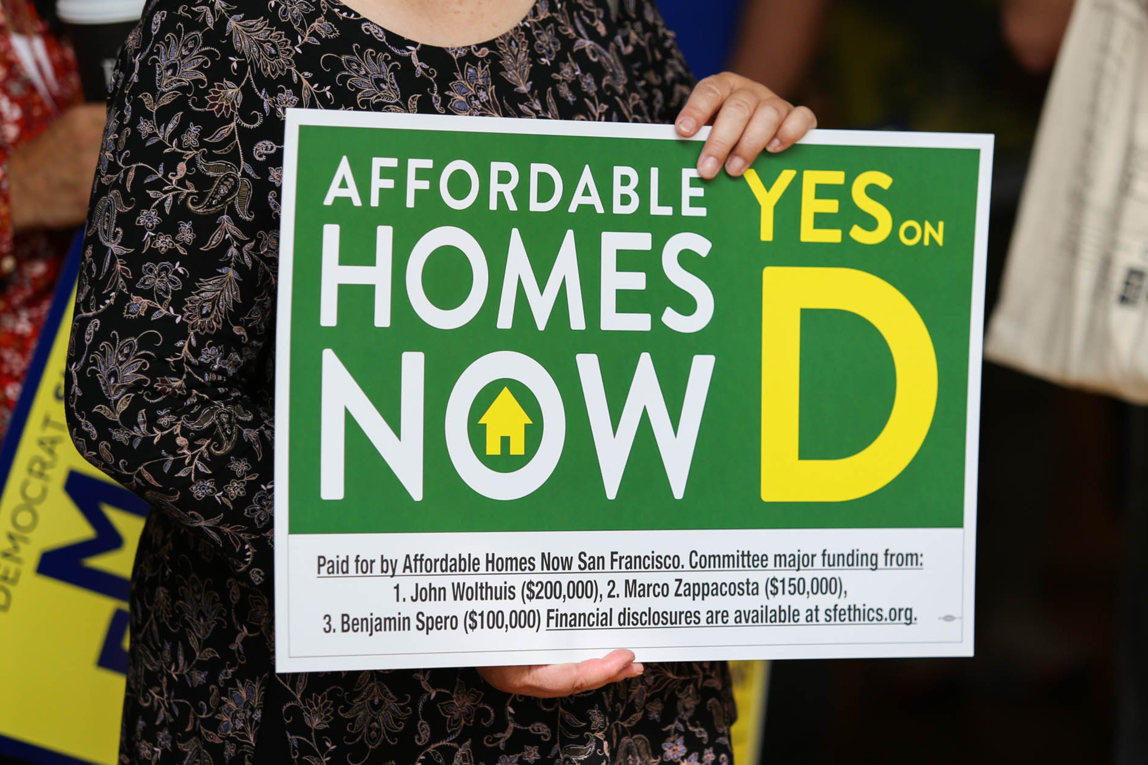 A person holds a green and yellow sign reading "Affordable Homes Now Yes on D" with funding details listed at the bottom.