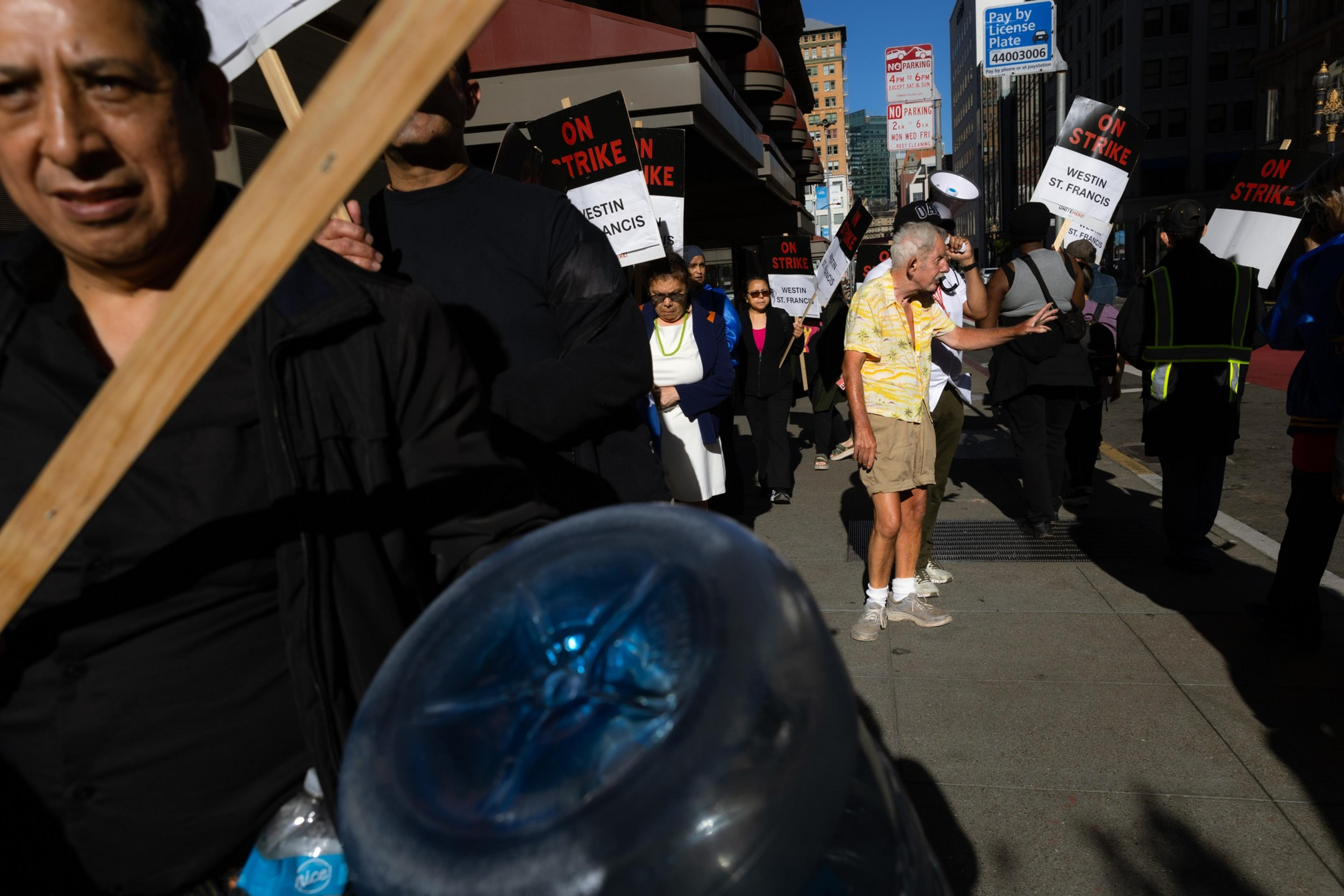 A group of people are marching on a sunny day, holding signs that read &quot;ON STRIKE&quot; with &quot;WESTIN ST. FRANCIS&quot; printed below. They appear determined and organized.
