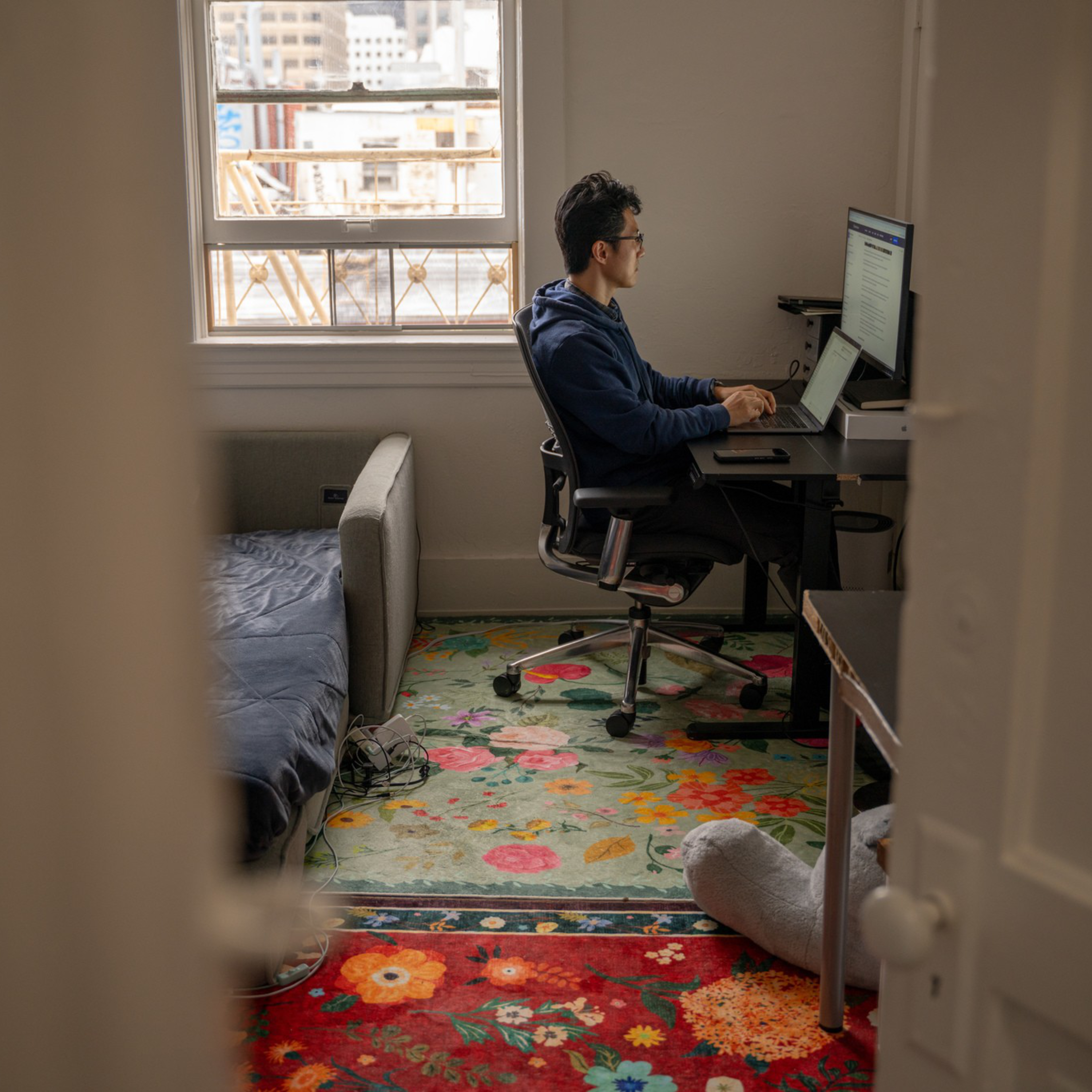 A person wearing glasses and a blue hoodie works on a computer at a desk in a small room with colorful floral rugs and a sofa beside a window.