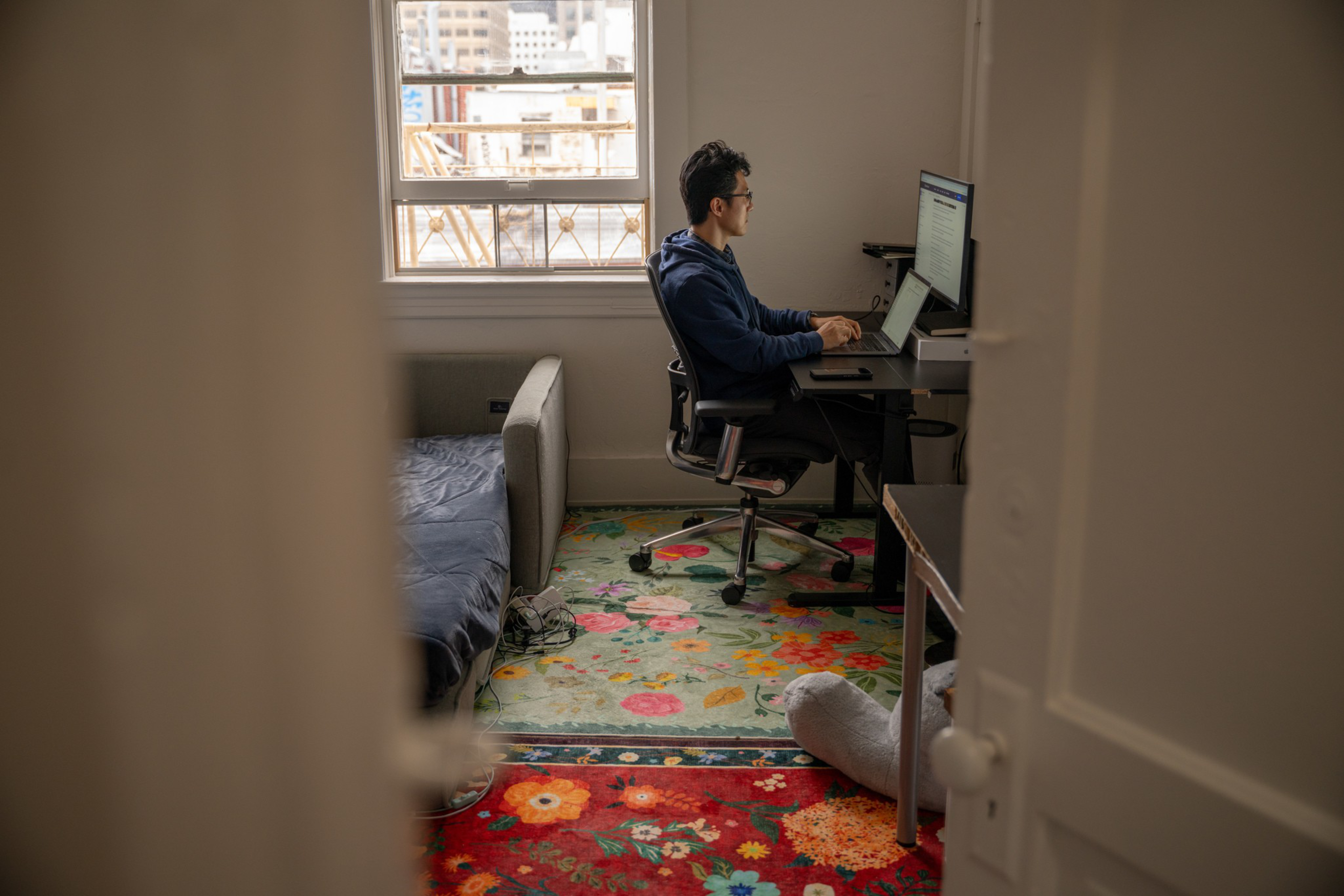 A person wearing glasses and a blue hoodie works on a computer at a desk in a small room with colorful floral rugs and a sofa beside a window.