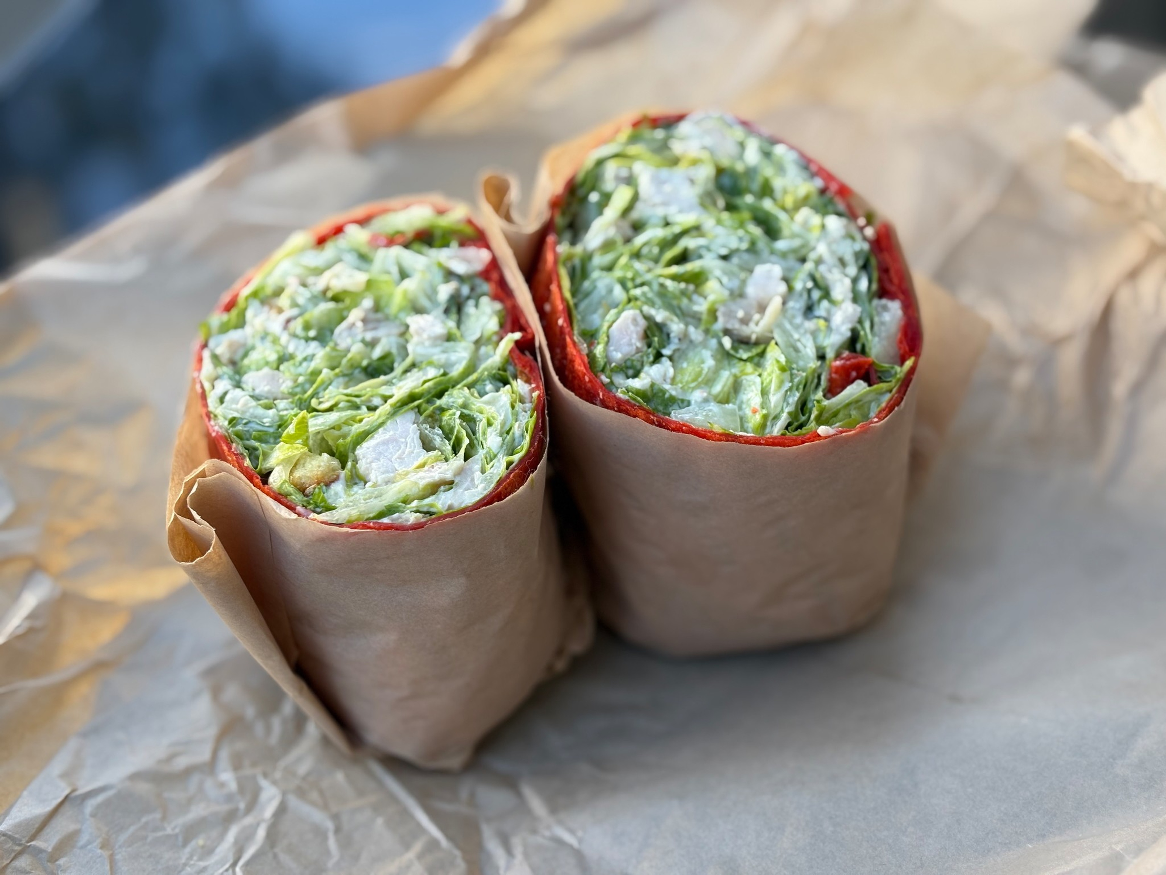 Two halves of a wrap with red tortilla, filled with chopped lettuce and pieces of chicken, wrapped in brown paper on crumpled paper surface.