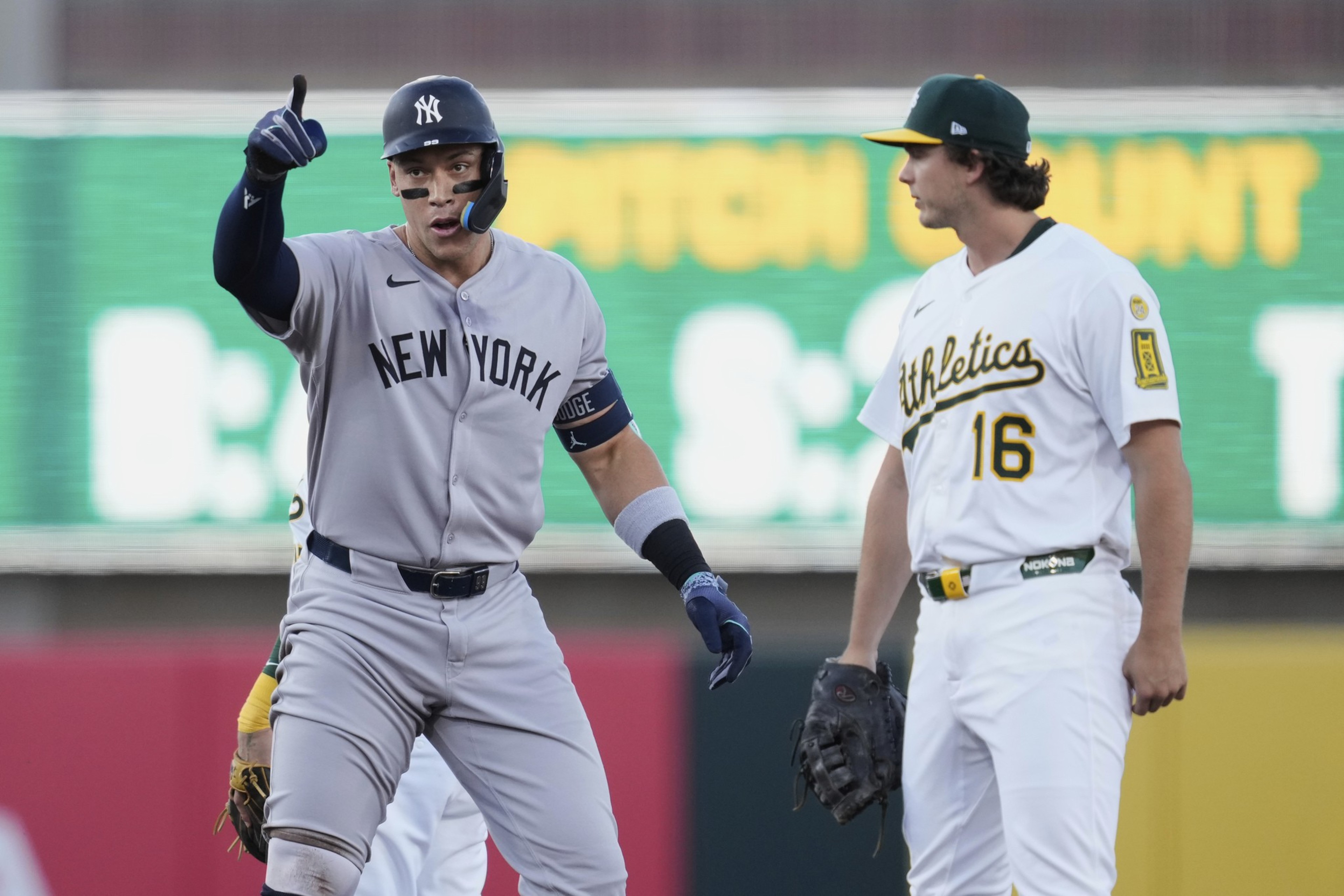Two baseball players are on the field. One, in a gray "New York" uniform, gestures with a thumbs up. The other, in a white "Athletics" uniform, observes.