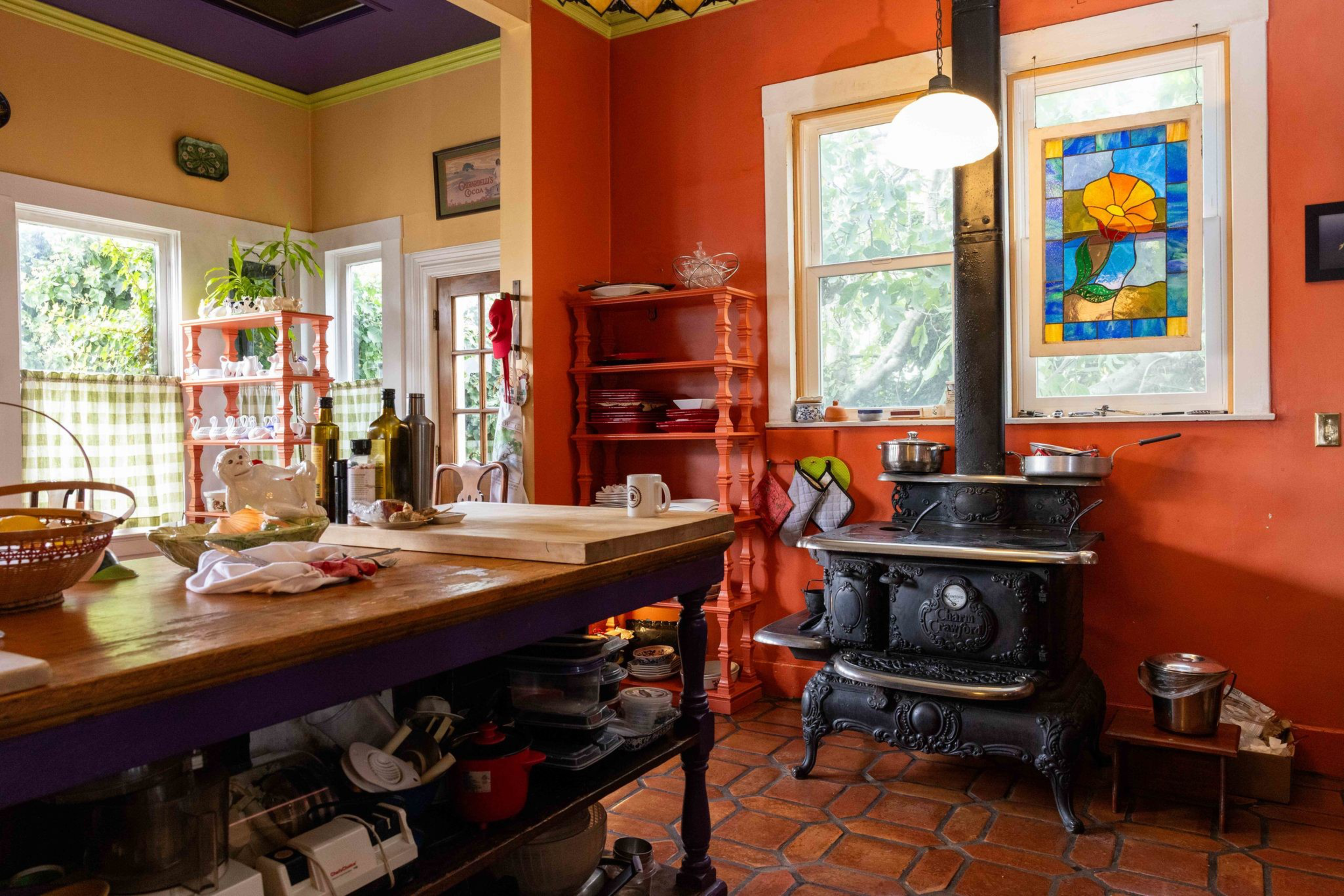 A cozy kitchen with orange walls, a black vintage stove, stained glass window, wooden shelves, and a table cluttered with kitchen items.