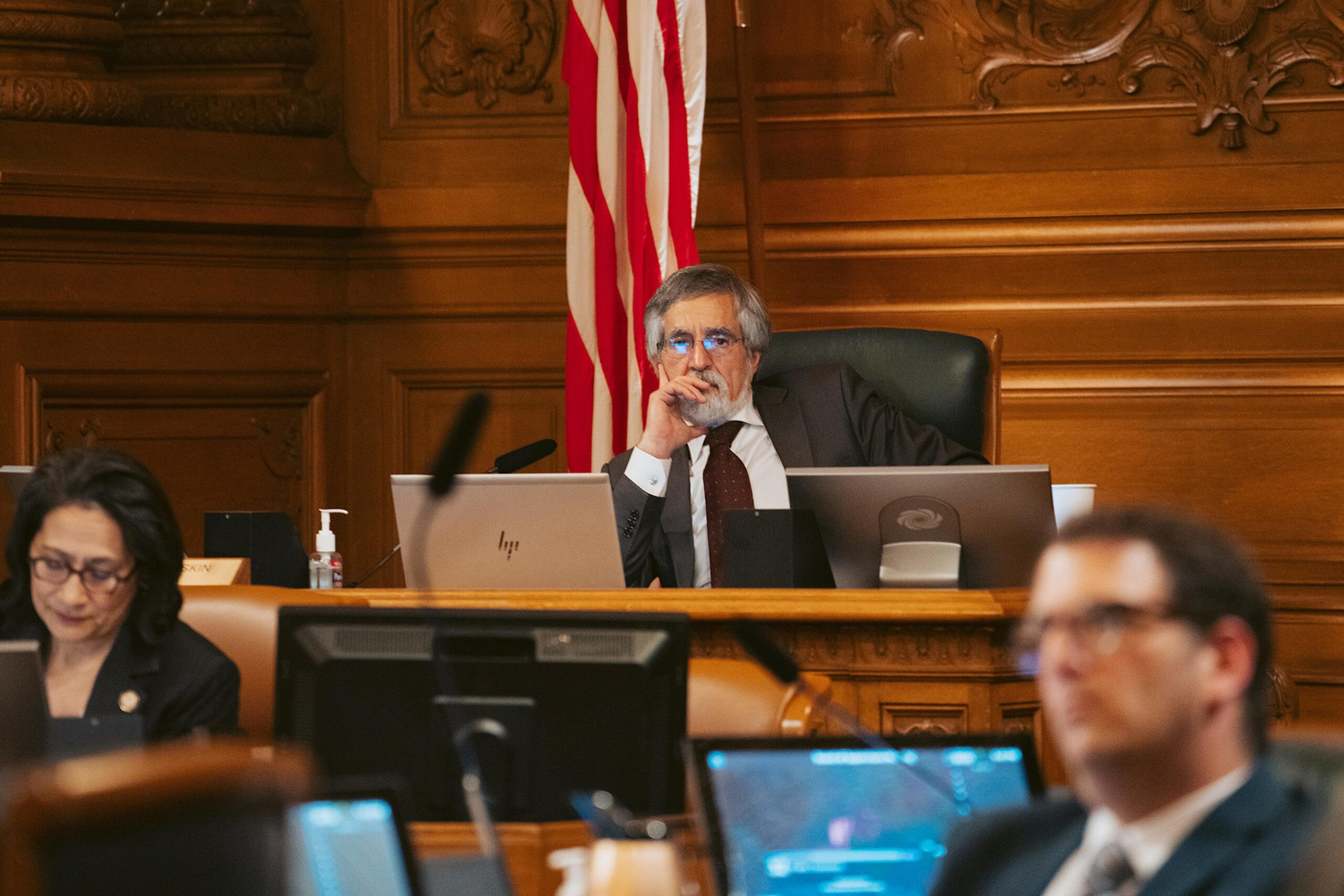 A man sits pensively behind a desk in a wood-paneled room with an American flag, surrounded by other focused individuals at desks with computers.