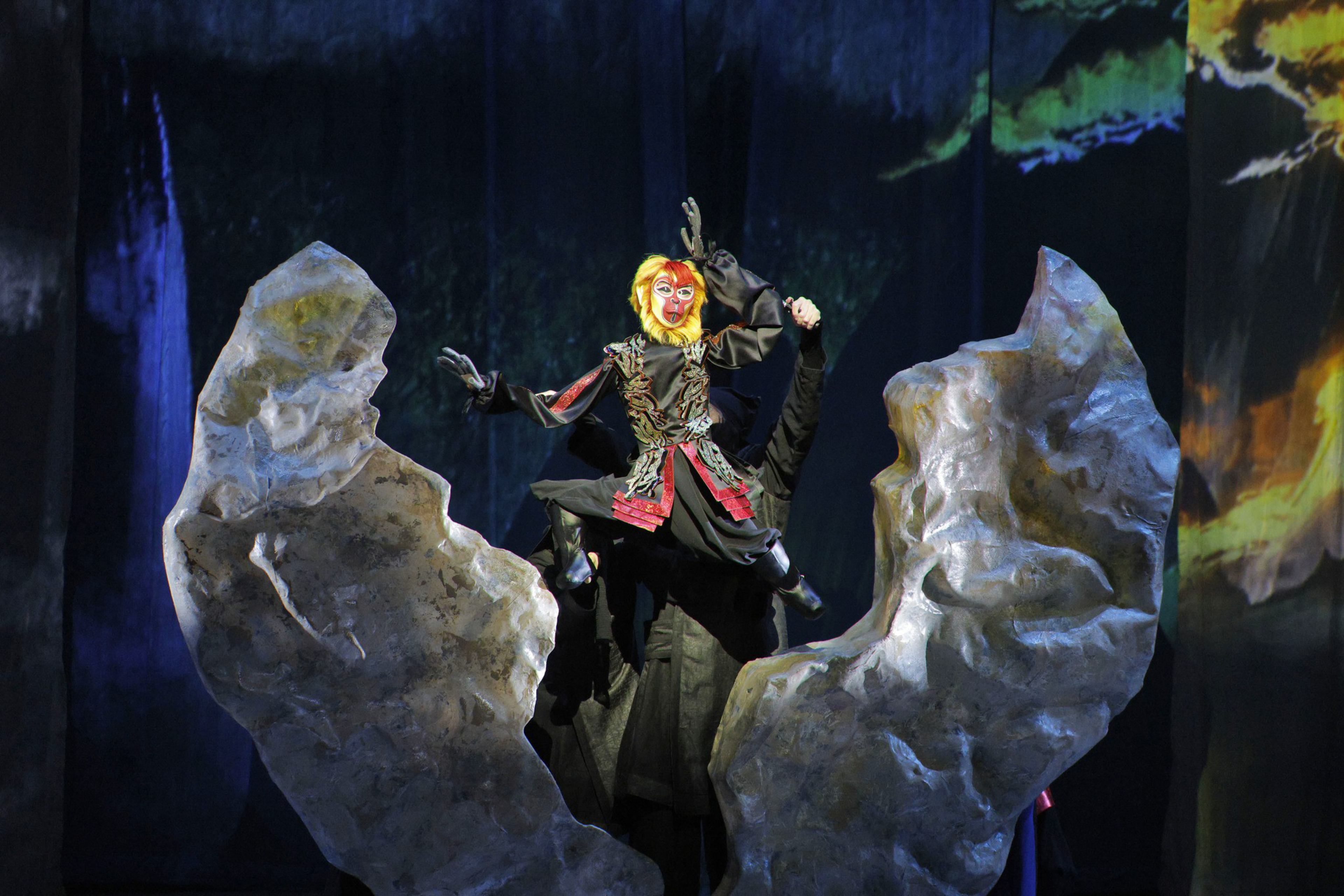 A performer in a colorful monkey costume leaps dramatically between two large, jagged rock props on a dark stage.