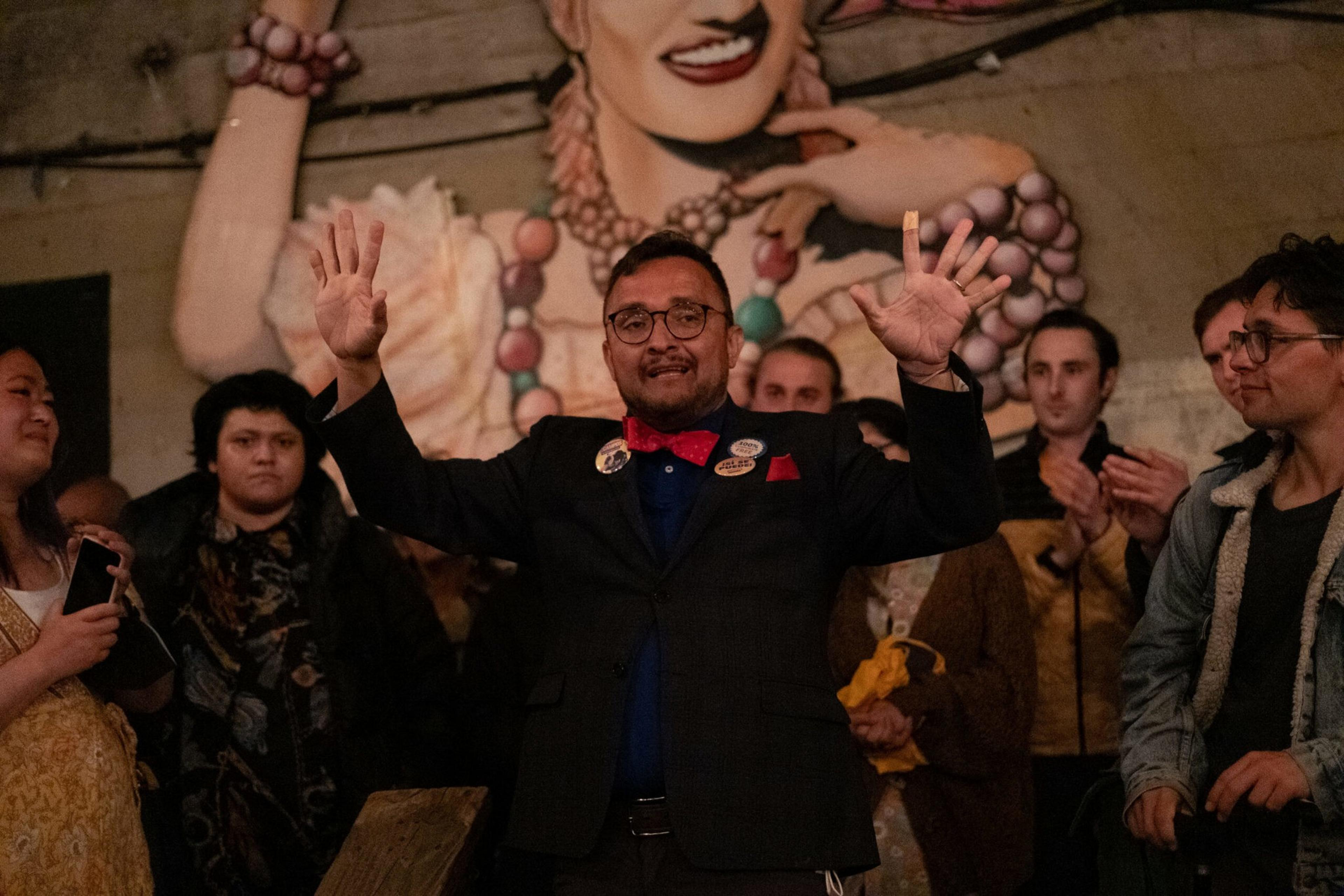 A man in a suit with a red bow tie raises both hands while people around him clap and smile, with a large mural of a smiling face behind them.