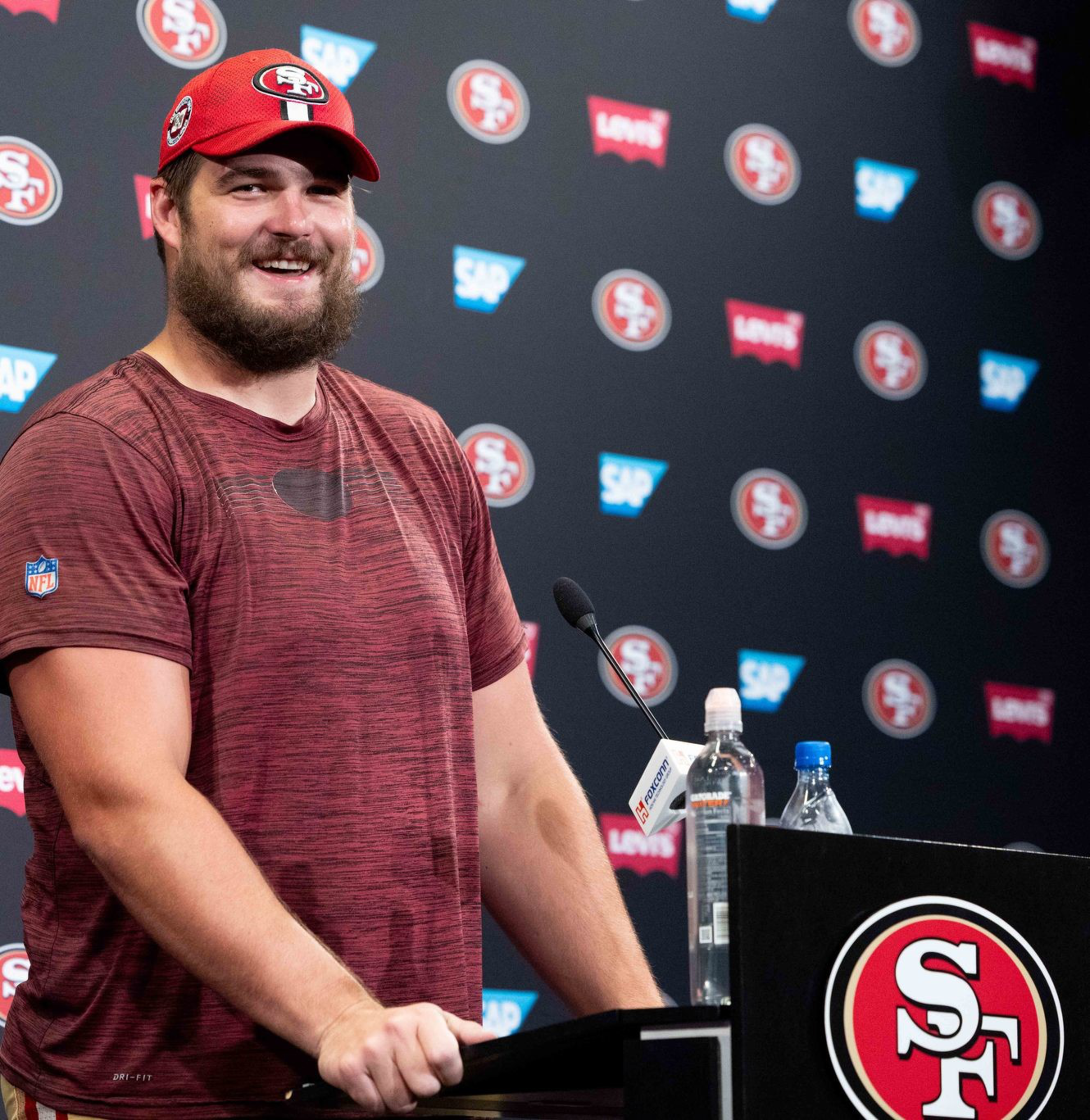 A smiling man in a red cap and maroon shirt stands at a podium decorated with the San Francisco 49ers logo.