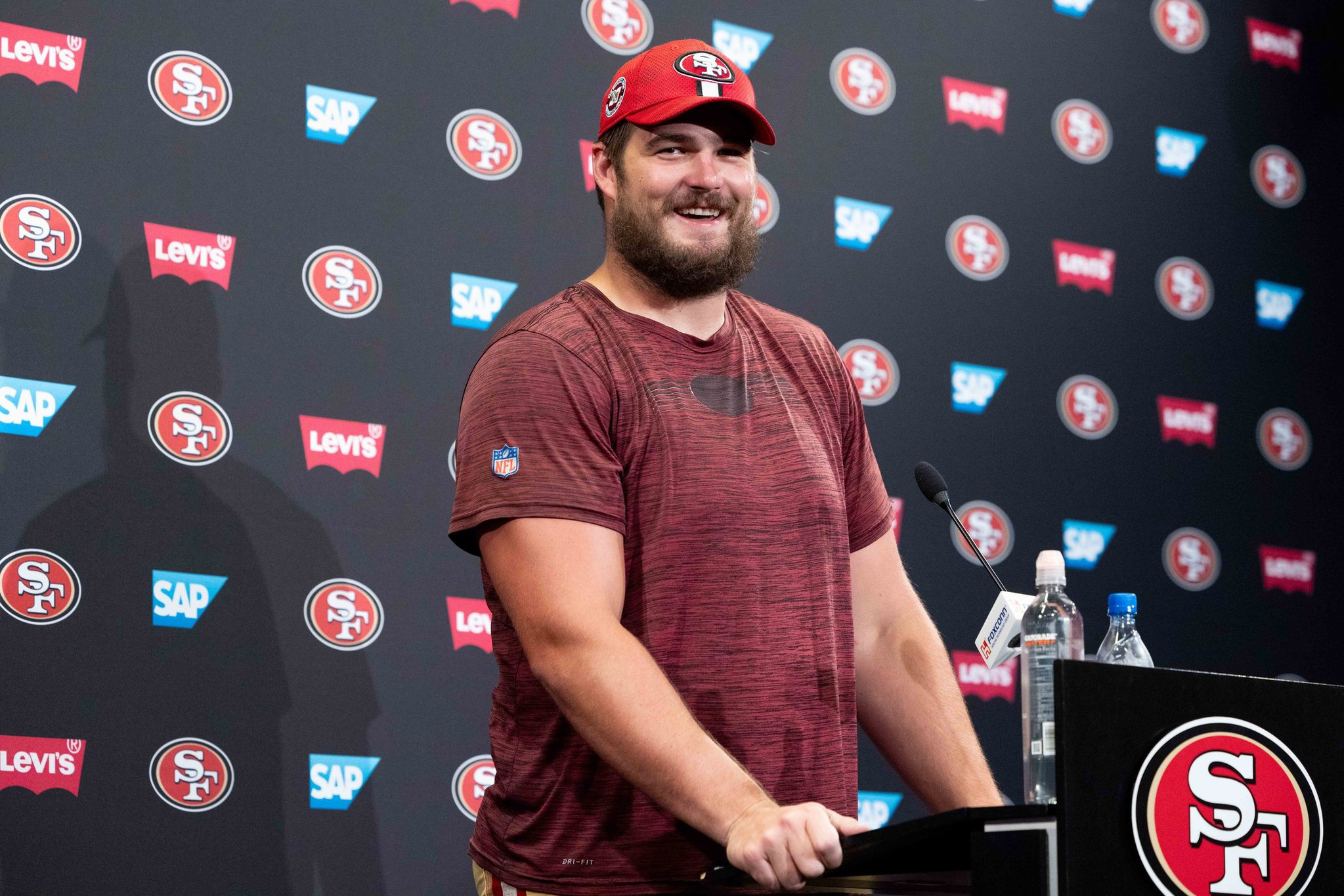 A smiling man in a red cap and maroon shirt stands at a podium decorated with the San Francisco 49ers logo.