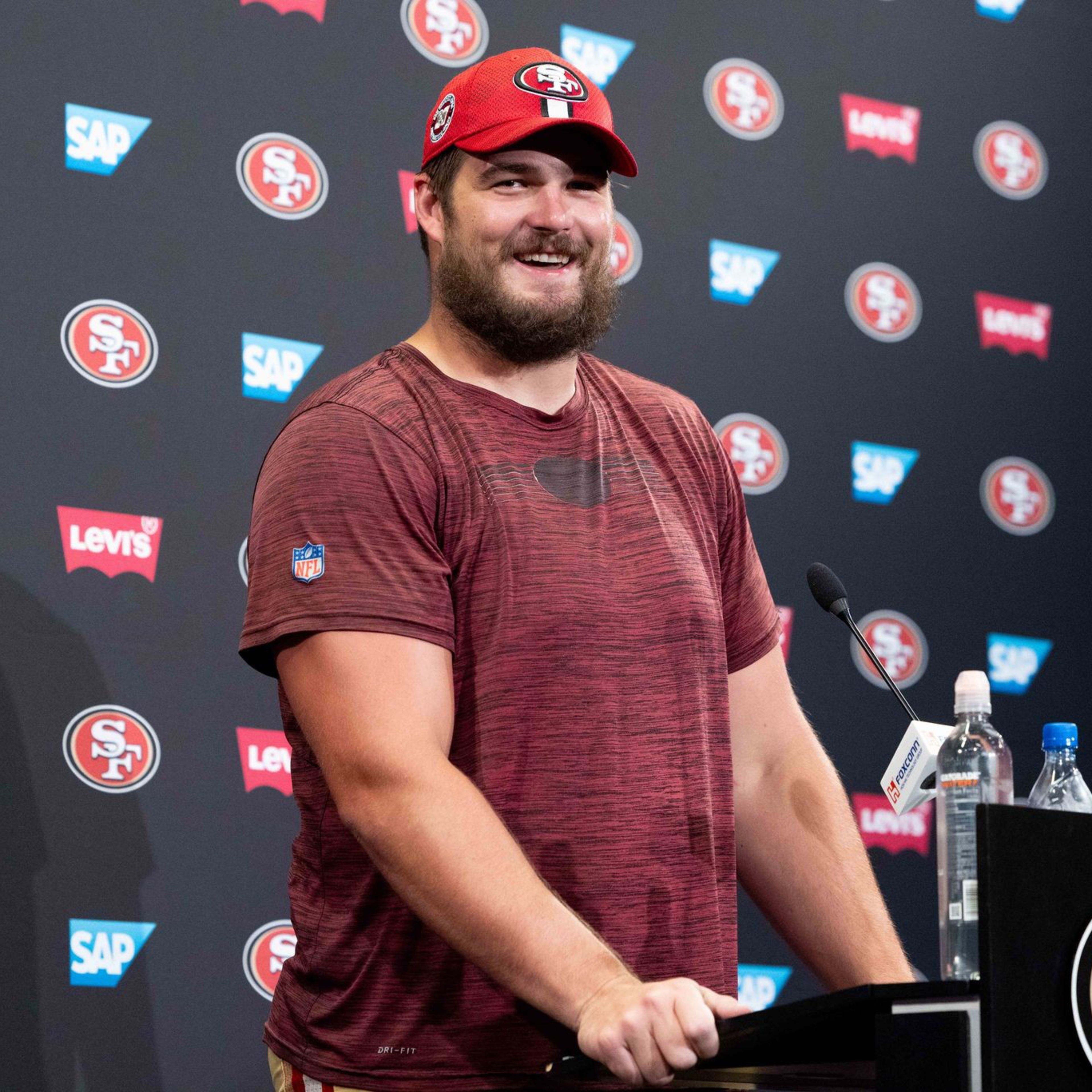 A smiling man in a red cap and maroon shirt stands at a podium decorated with the San Francisco 49ers logo.