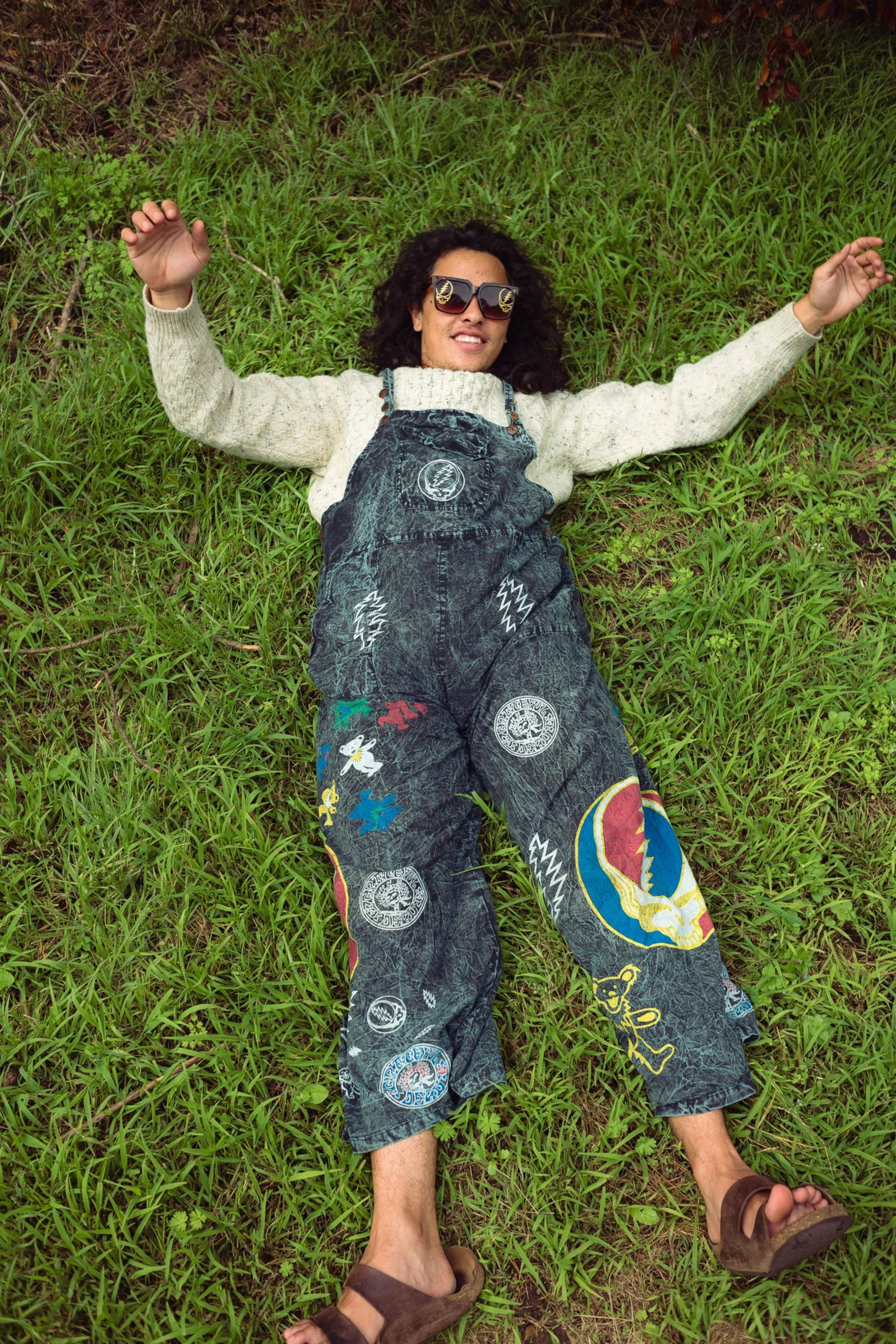 A person with curly hair wearing sunglasses, a light sweater, and colorful overalls lies relaxed on green grass with arms stretched out.