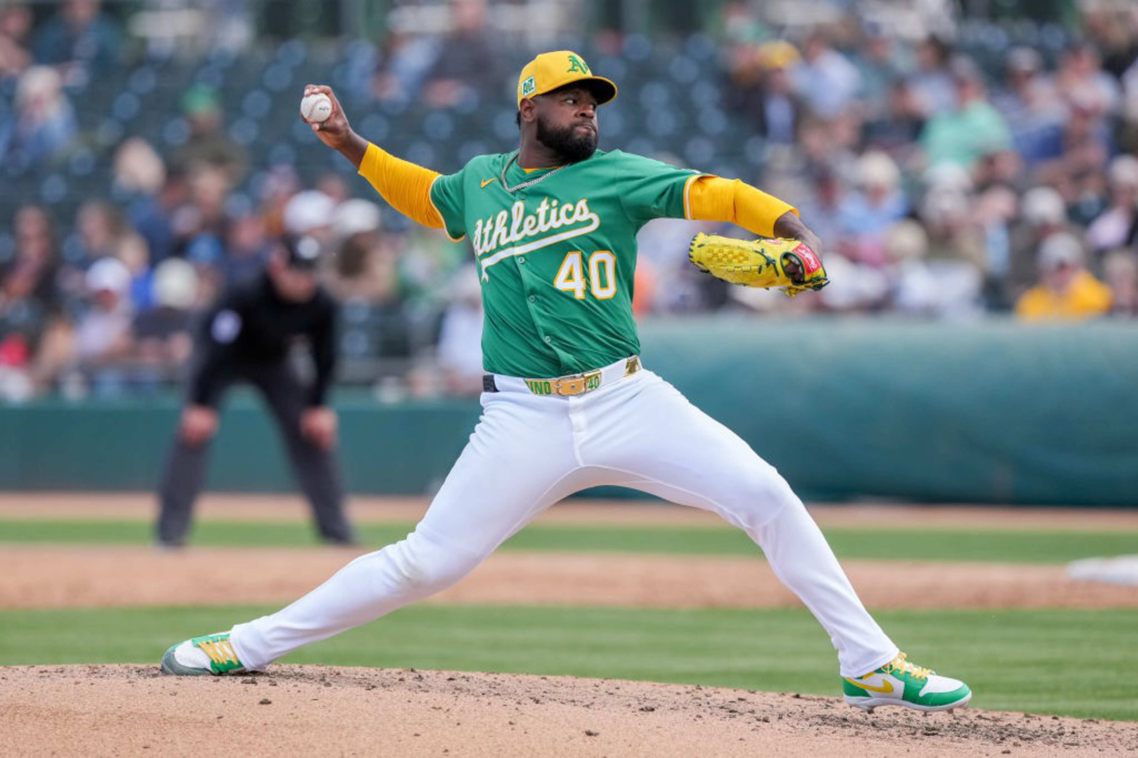 A baseball pitcher in a green jersey is mid-throw on a sunny field, wearing yellow gloves and cleats. An umpire and spectators are visible in the background.