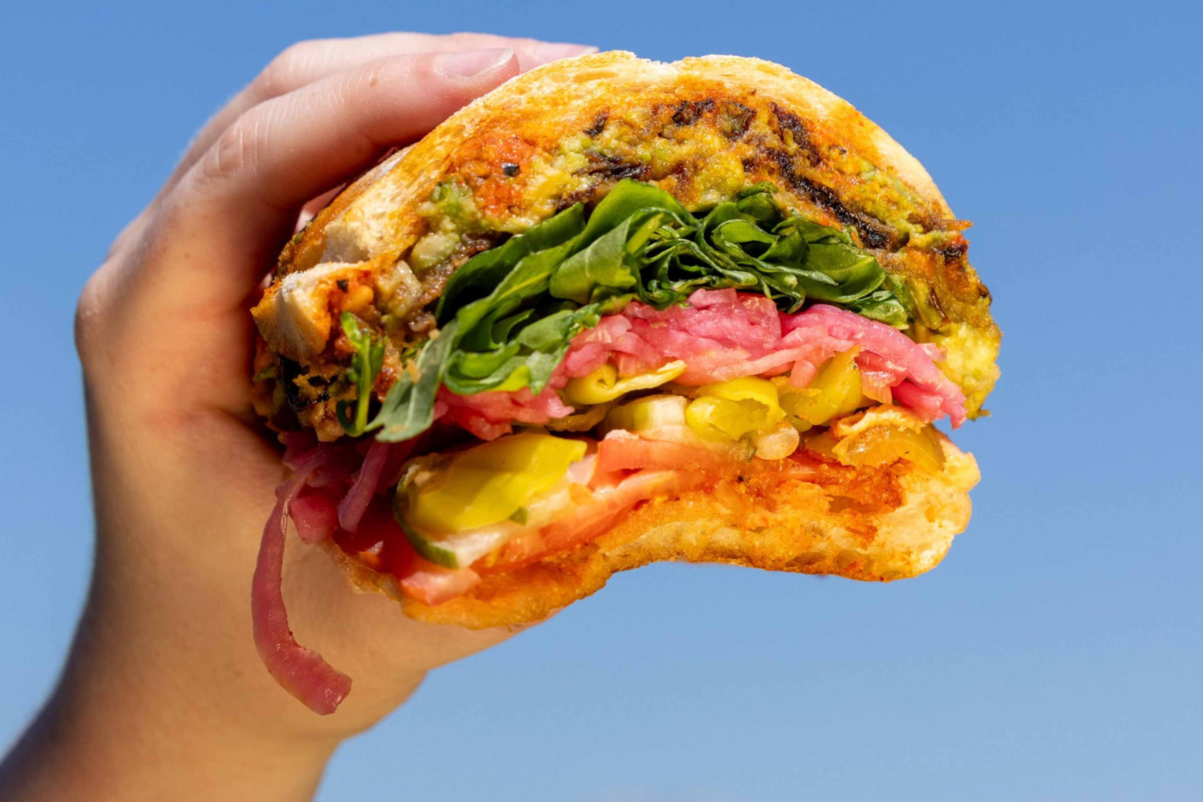 A hand holds a partially eaten sandwich with layers of meats, greens, and vegetables against a clear blue sky background.
