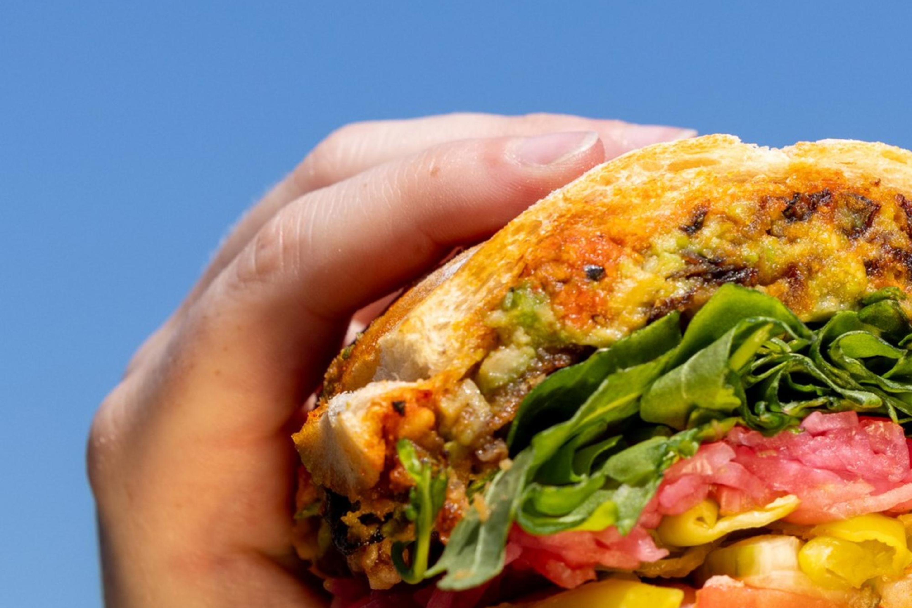 A hand holds a partially eaten sandwich with layers of meats, greens, and vegetables against a clear blue sky background.