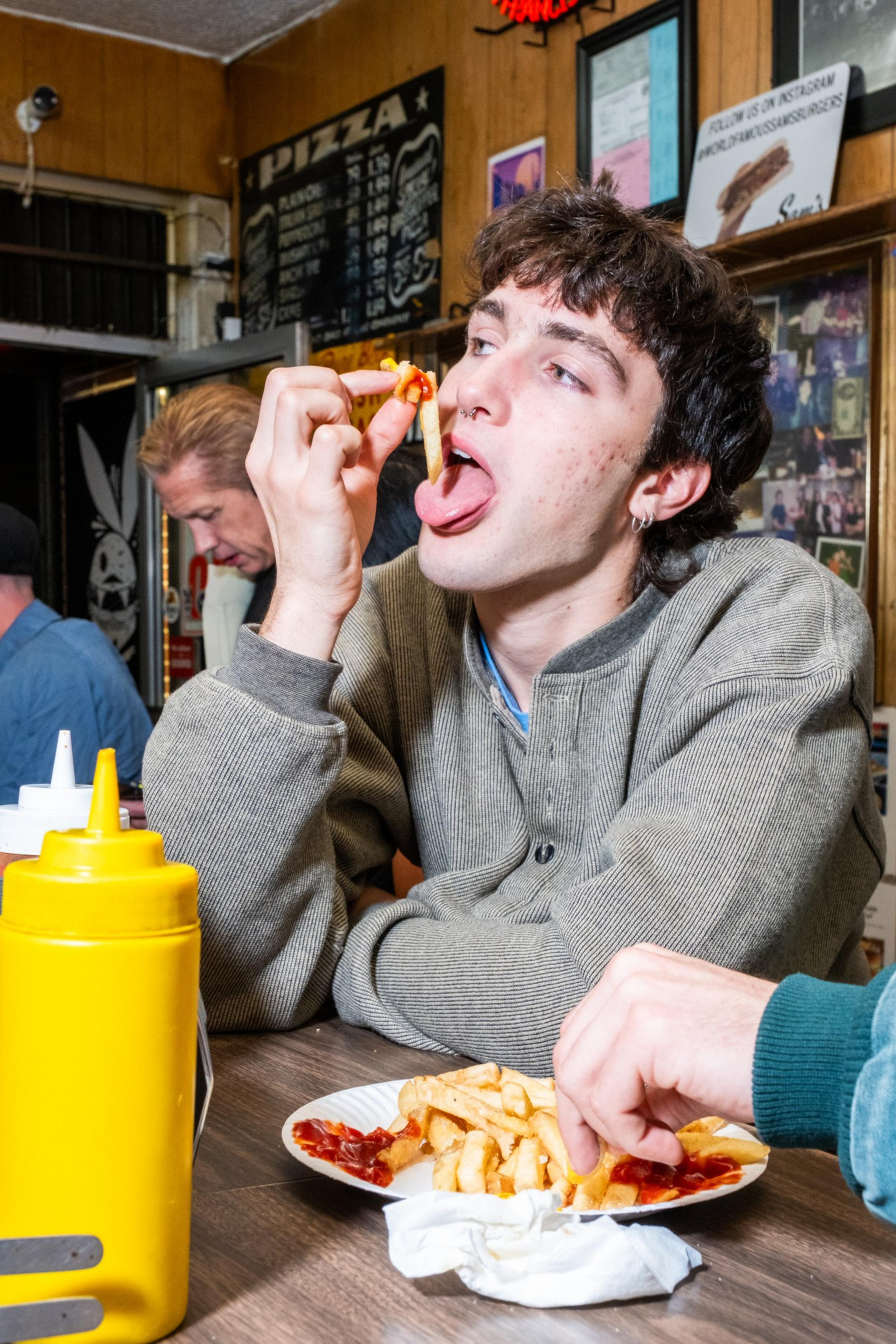 A person with short hair and earrings is holding a french fry with tomato sauce near their mouth in a diner, with a man blurred in the background.