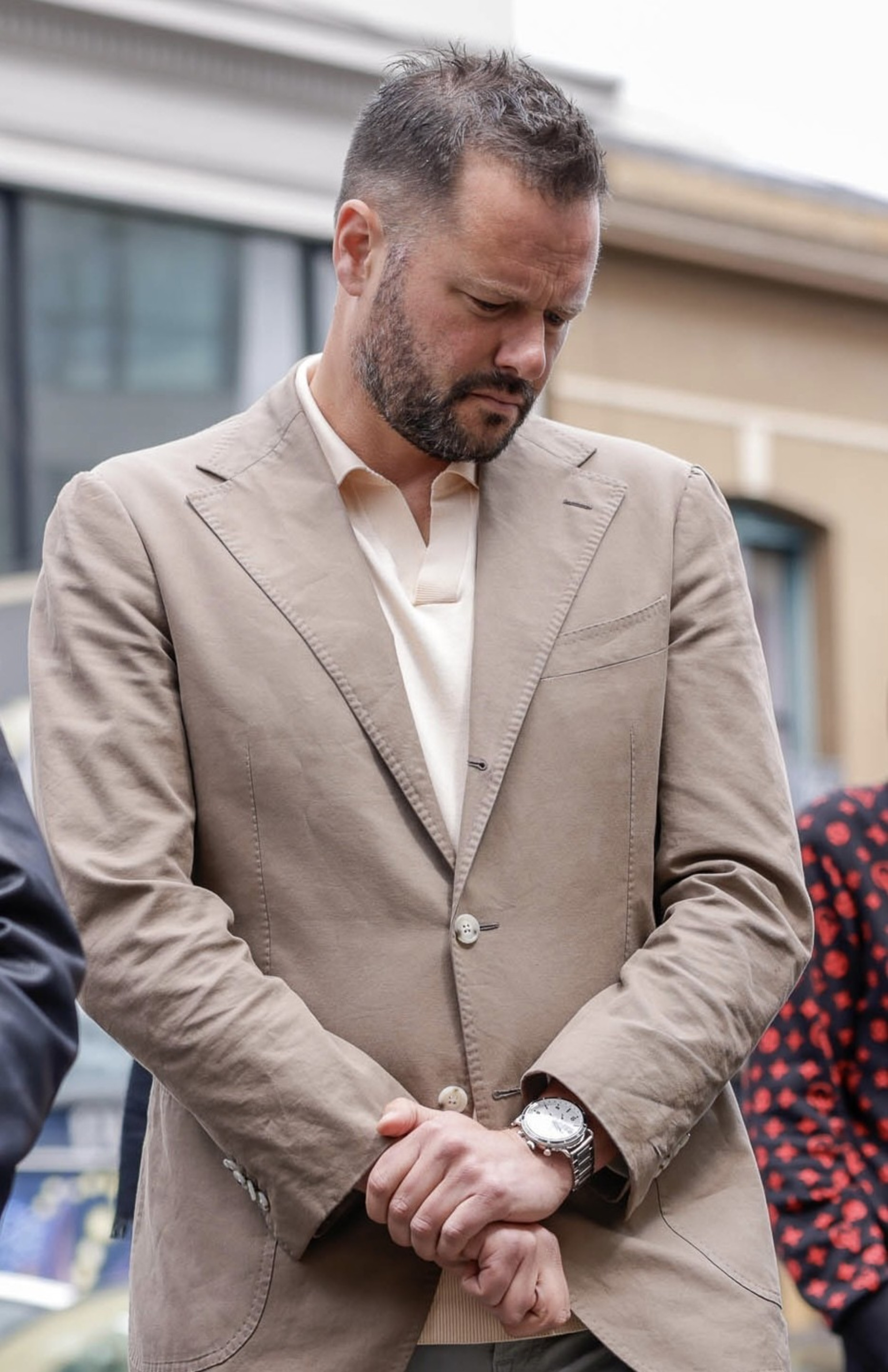 A man with short hair and a beard is wearing a beige blazer over a cream shirt. He looks down thoughtfully, with his hands clasped in front of him.