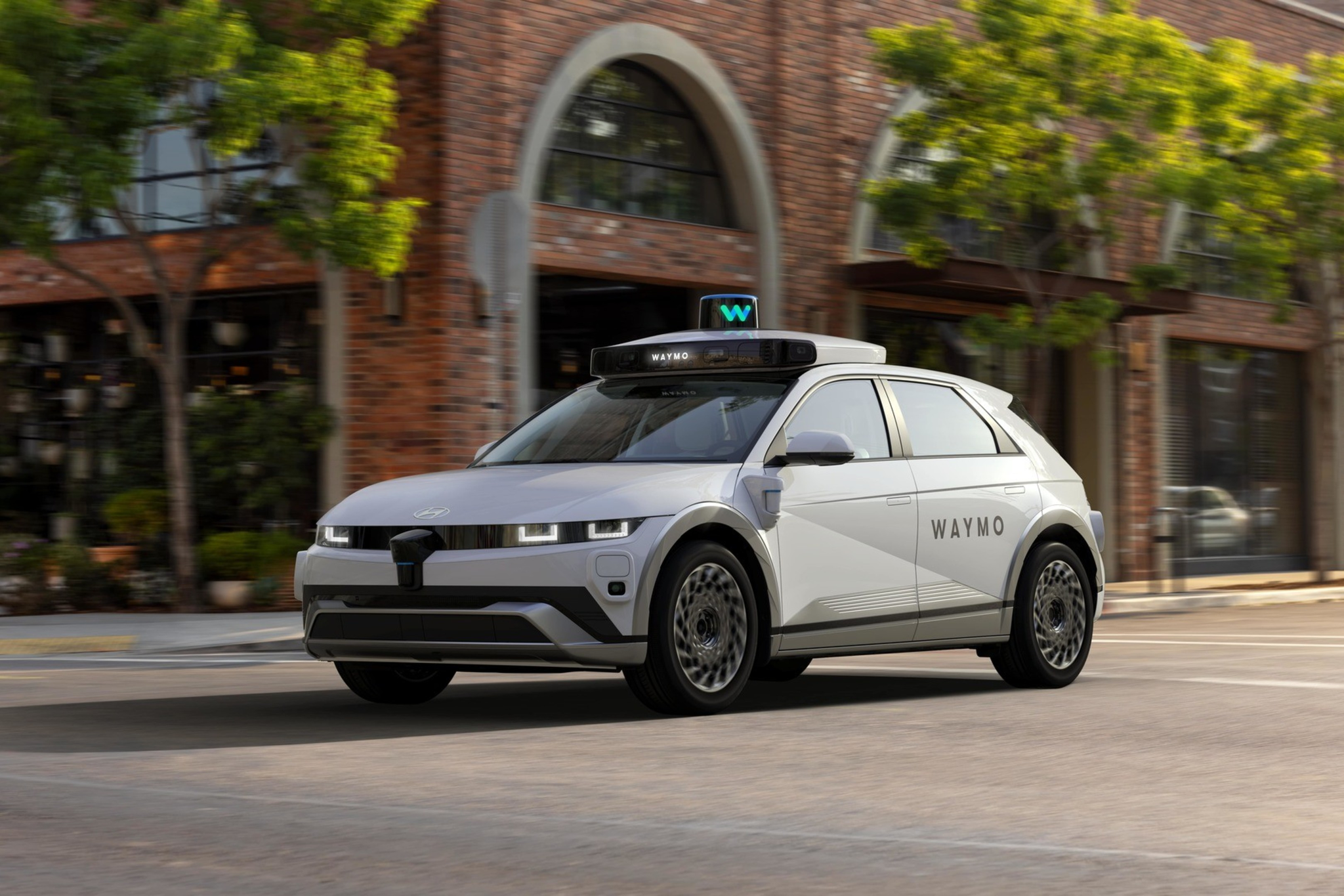 A white Waymo self-driving car drives on a city street with brick buildings and trees in the background.