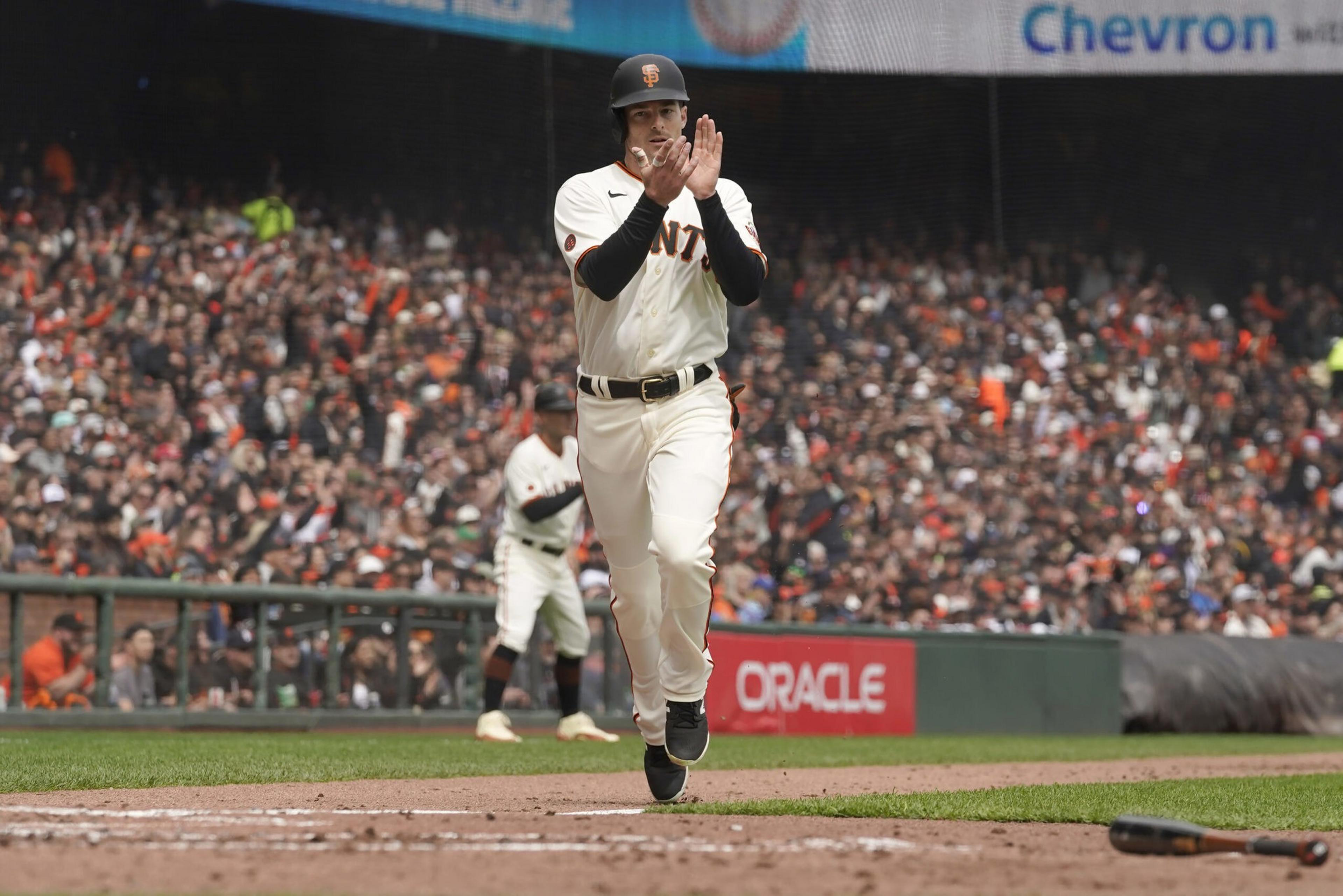 A San Francisco Giants player is running and clapping near home plate during a game with a large crowd in the stands behind him.