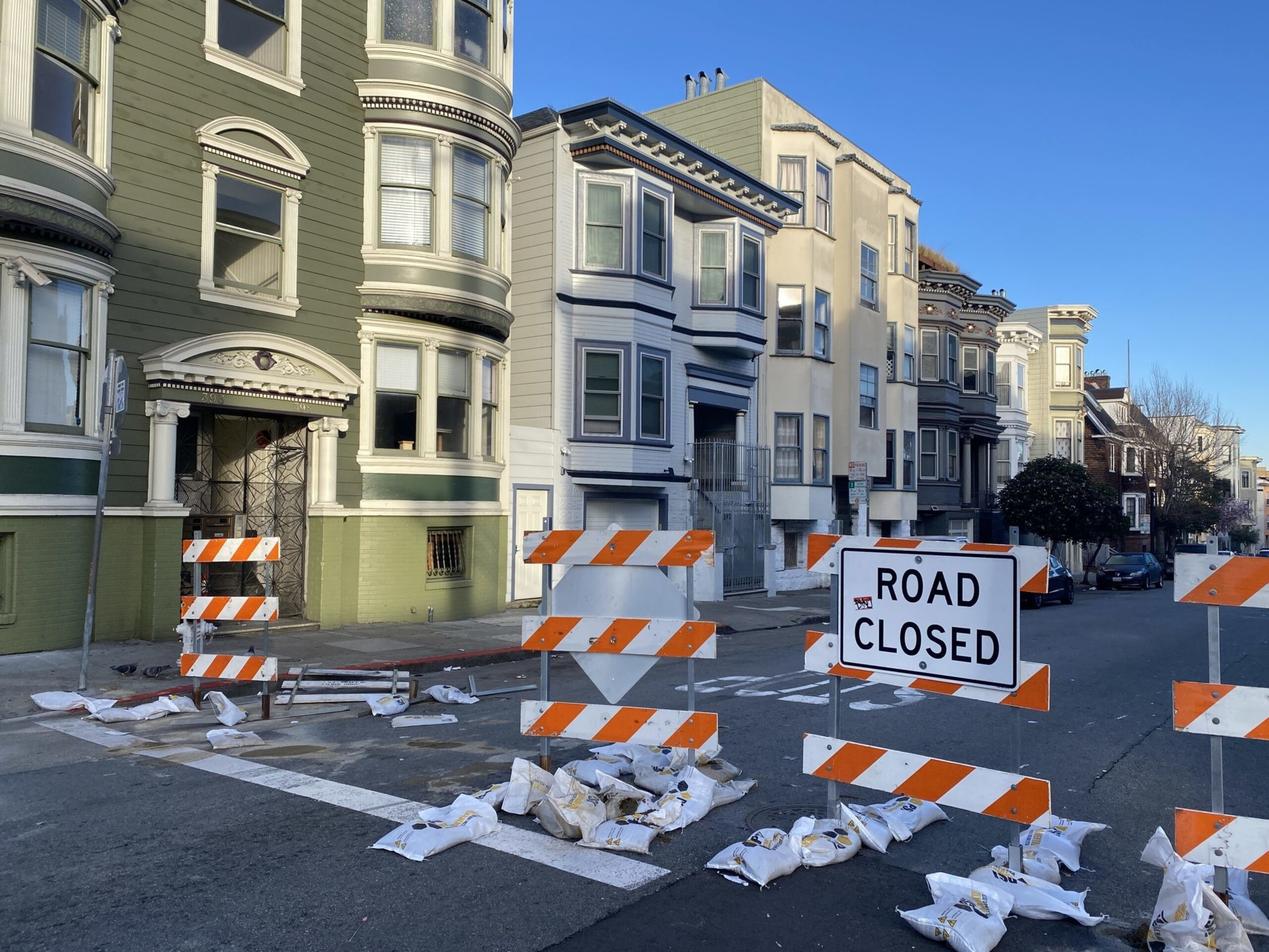 A street lined with multi-story houses is blocked by orange and white barricades and a "ROAD CLOSED" sign with sandbags at their bases.