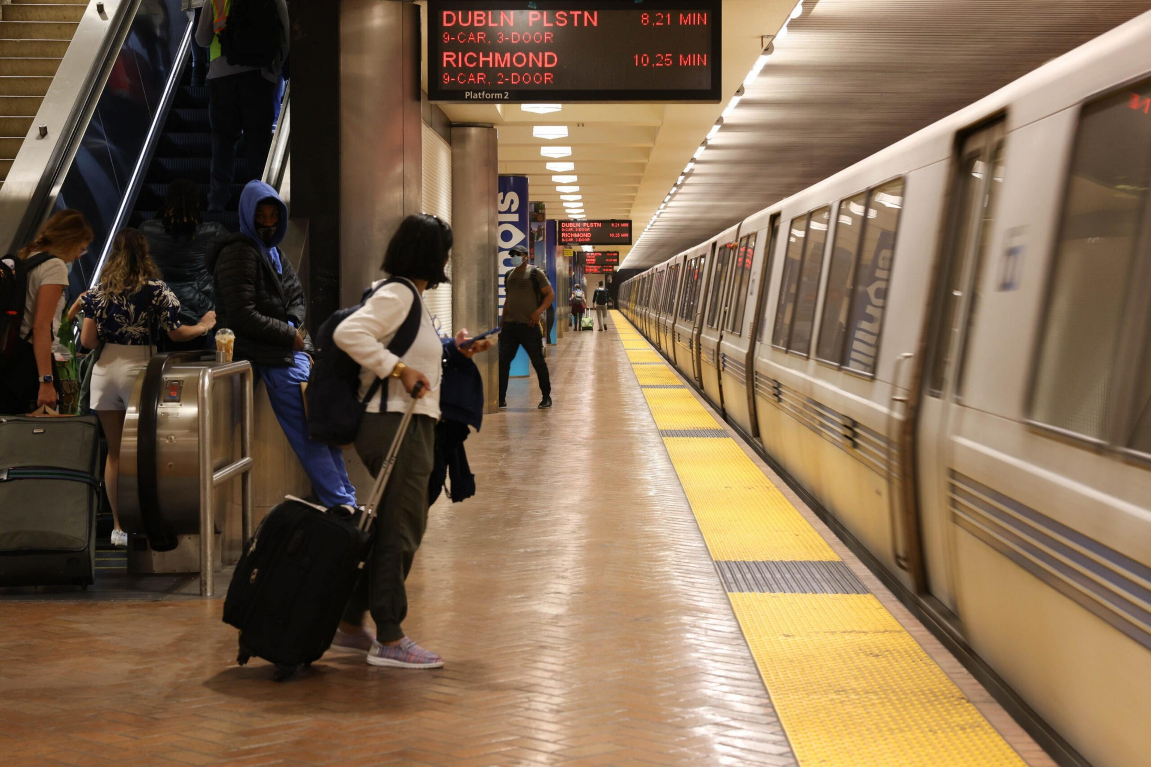 Commuters wait on a subway platform as a train arrives, with an escalator and digital sign displaying train times in the background.