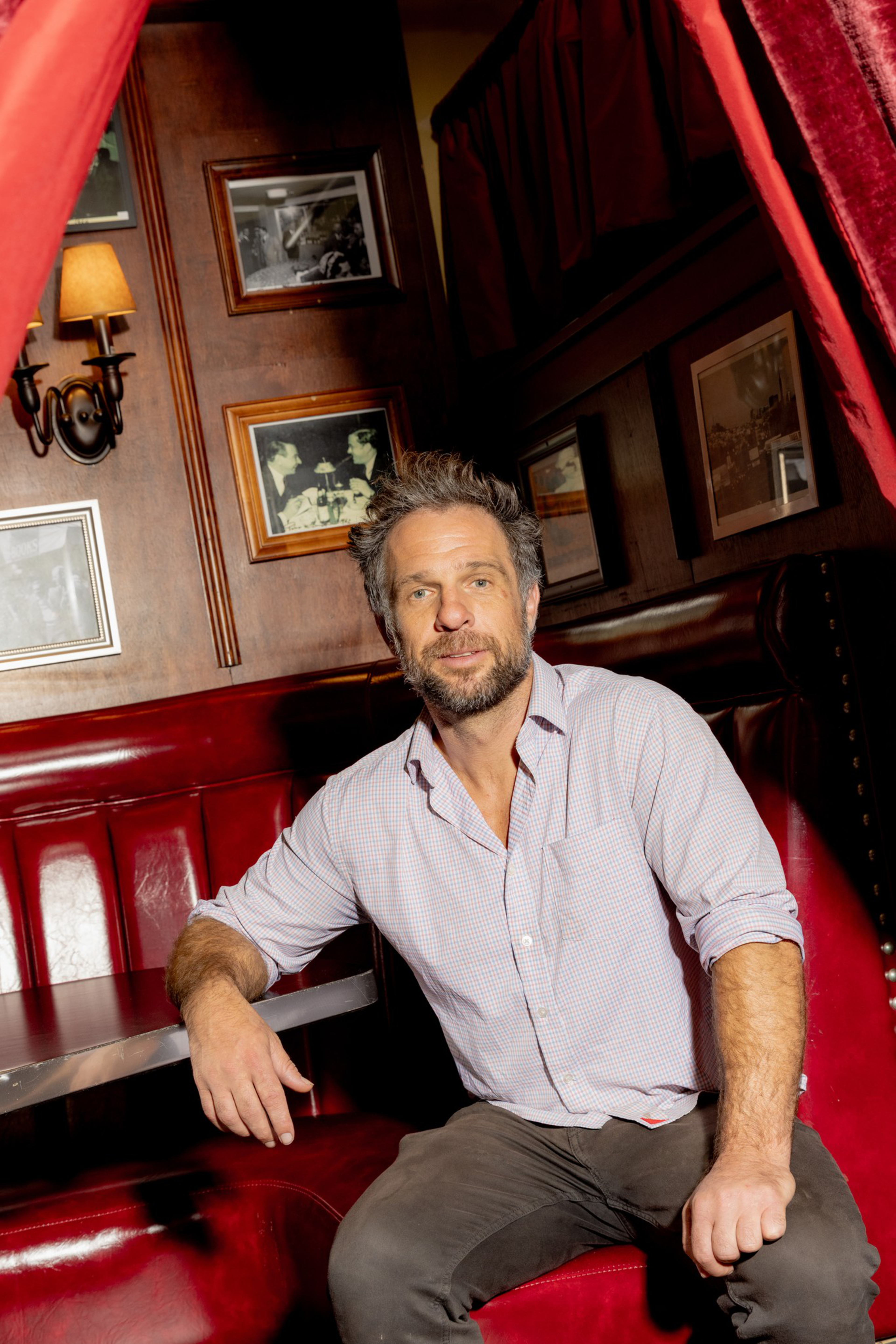 A man with a beard and casual clothes sits relaxed in a red leather booth inside a dimly lit, vintage-style restaurant or bar.