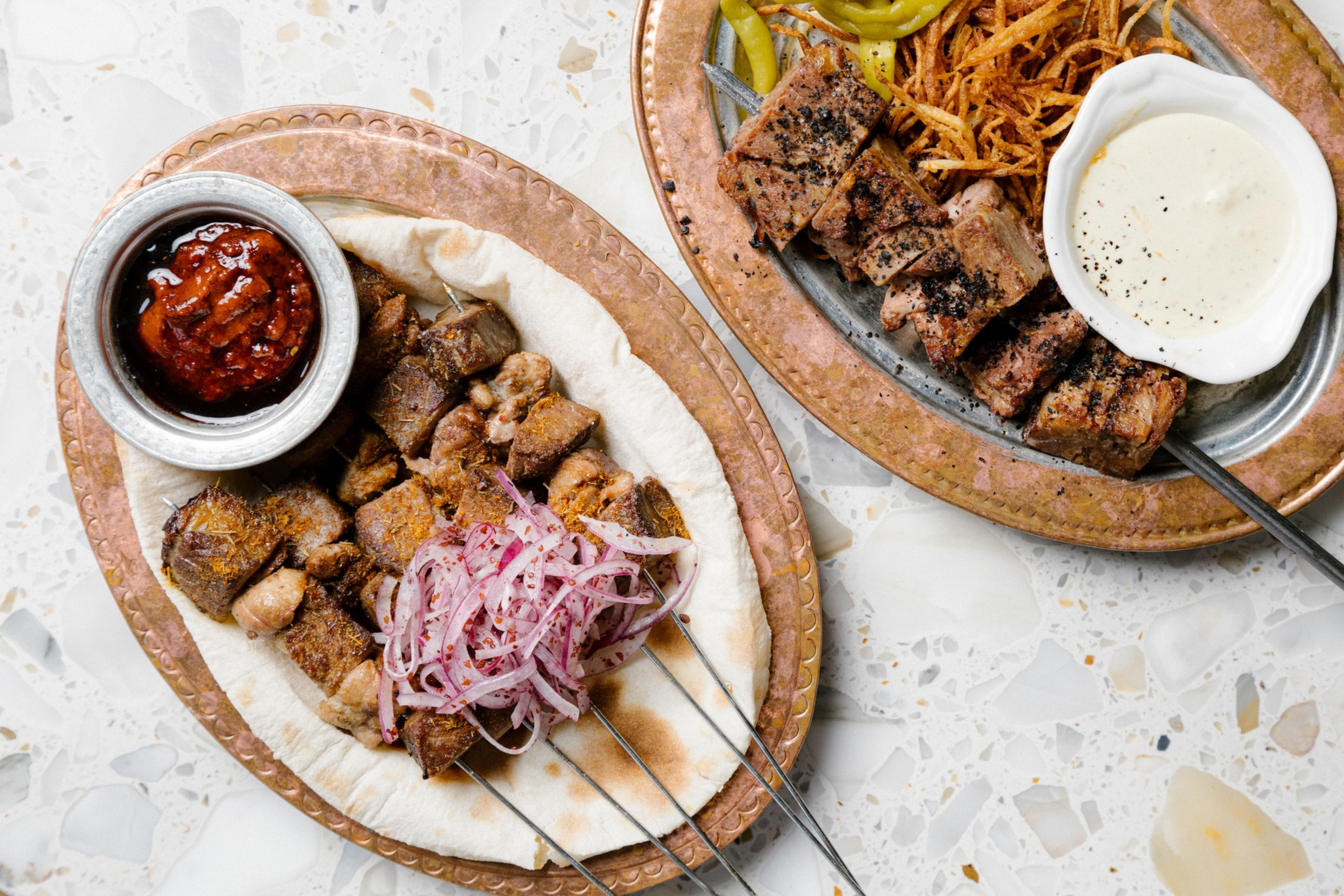 A close up photo of 2 dishes that include meat skewers, pita bread, peppers, and red and white dipping sauces on a marble table.