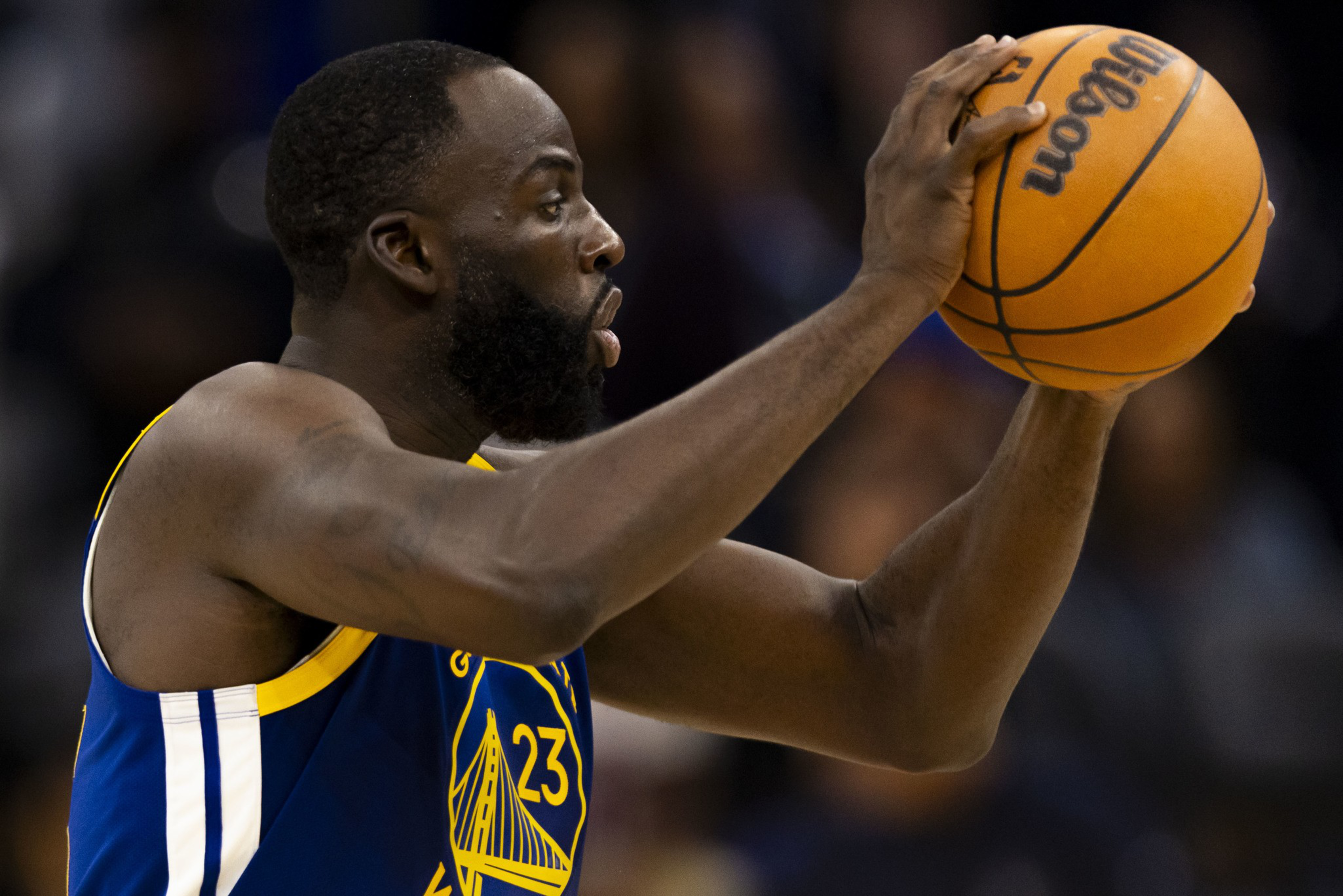 A basketball player wearing a blue and yellow Golden State Warriors jersey holds a basketball with both hands, focused on his next move.