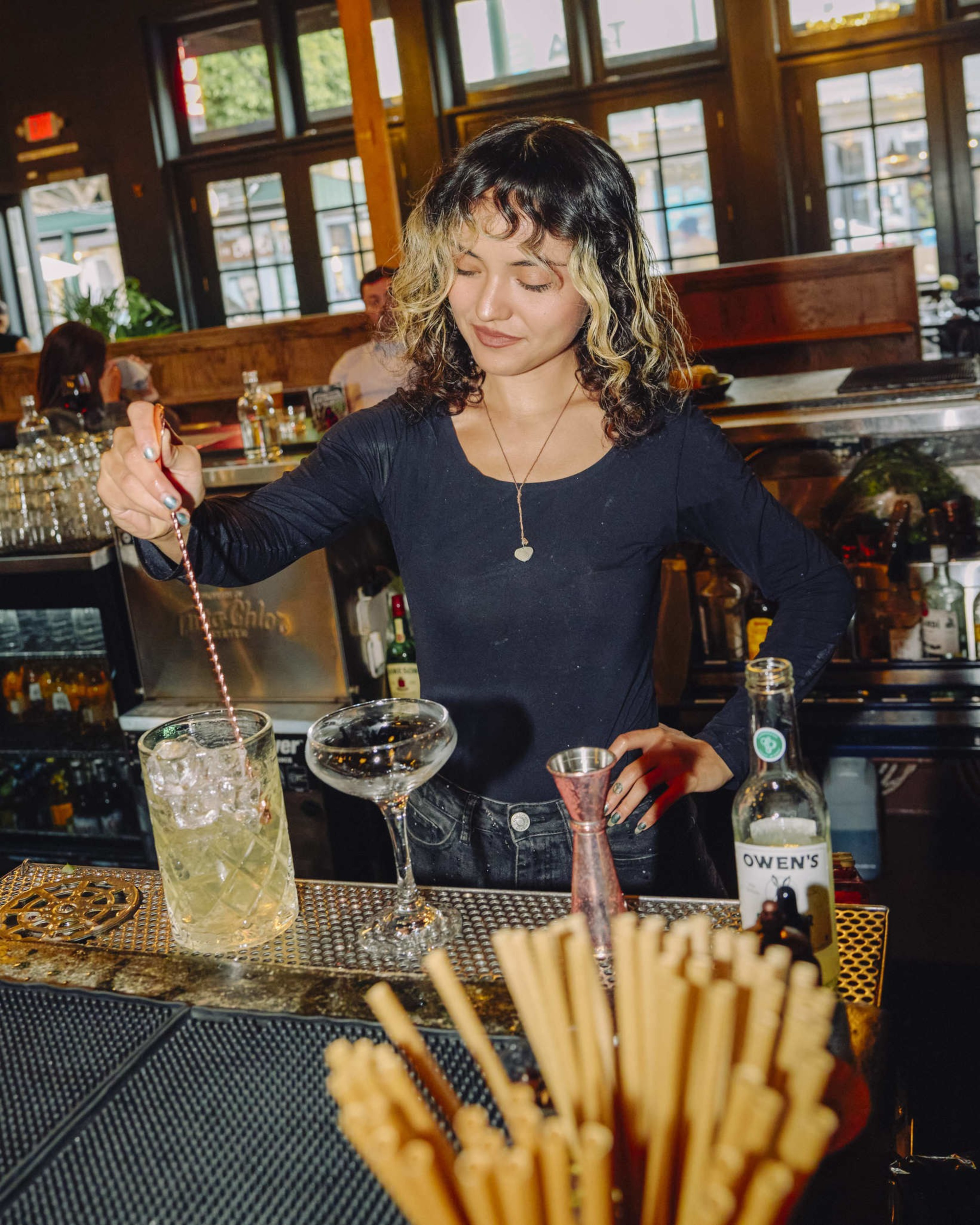 A woman with curly hair and light streaks stirs a drink with ice behind a bar, surrounded by bottles, a cocktail glass, and a container of straws.