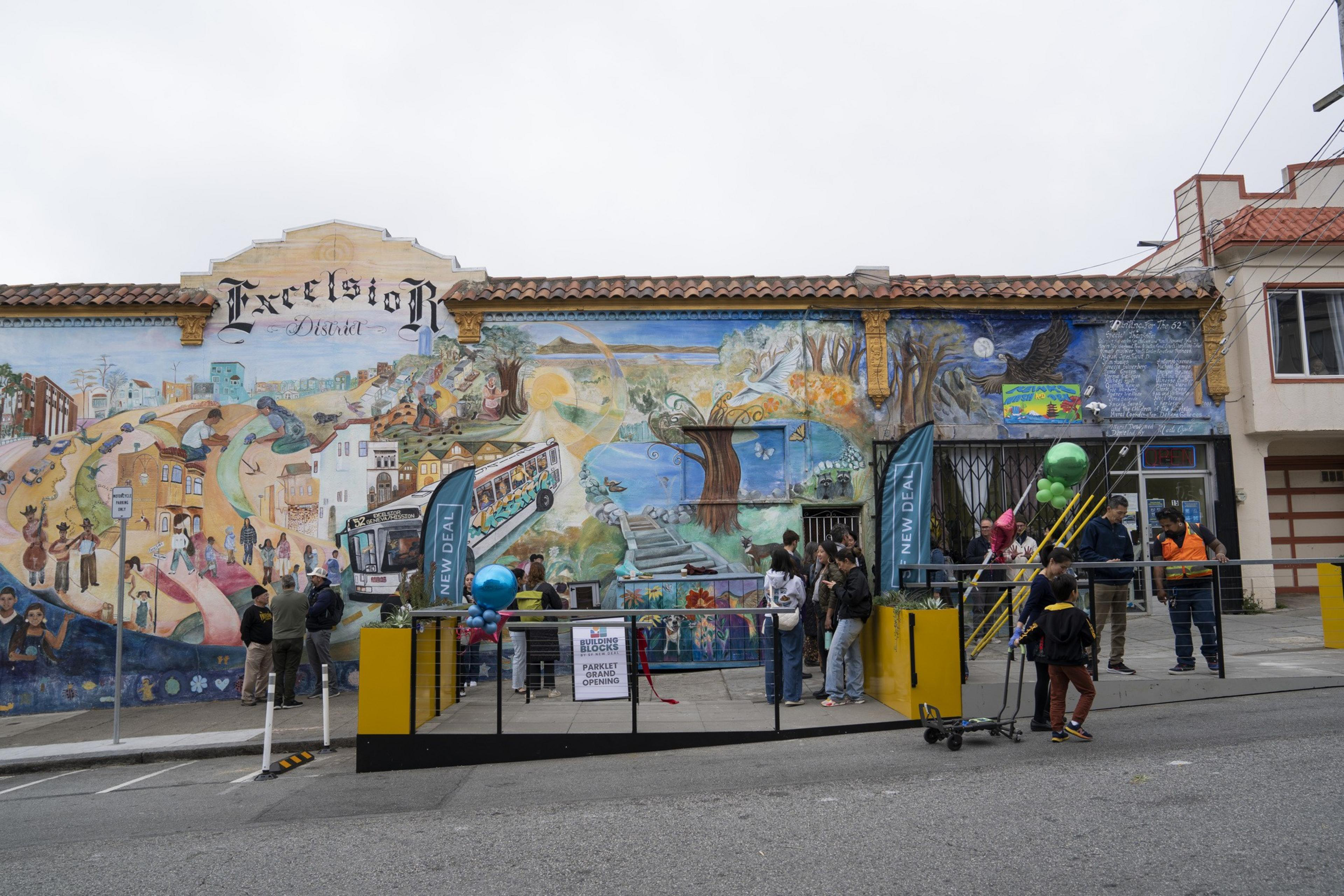 People are gathered in front of a colorful mural depicting urban and natural scenes, with a sign saying "Excelsior District." Balloons and flags add festivity.