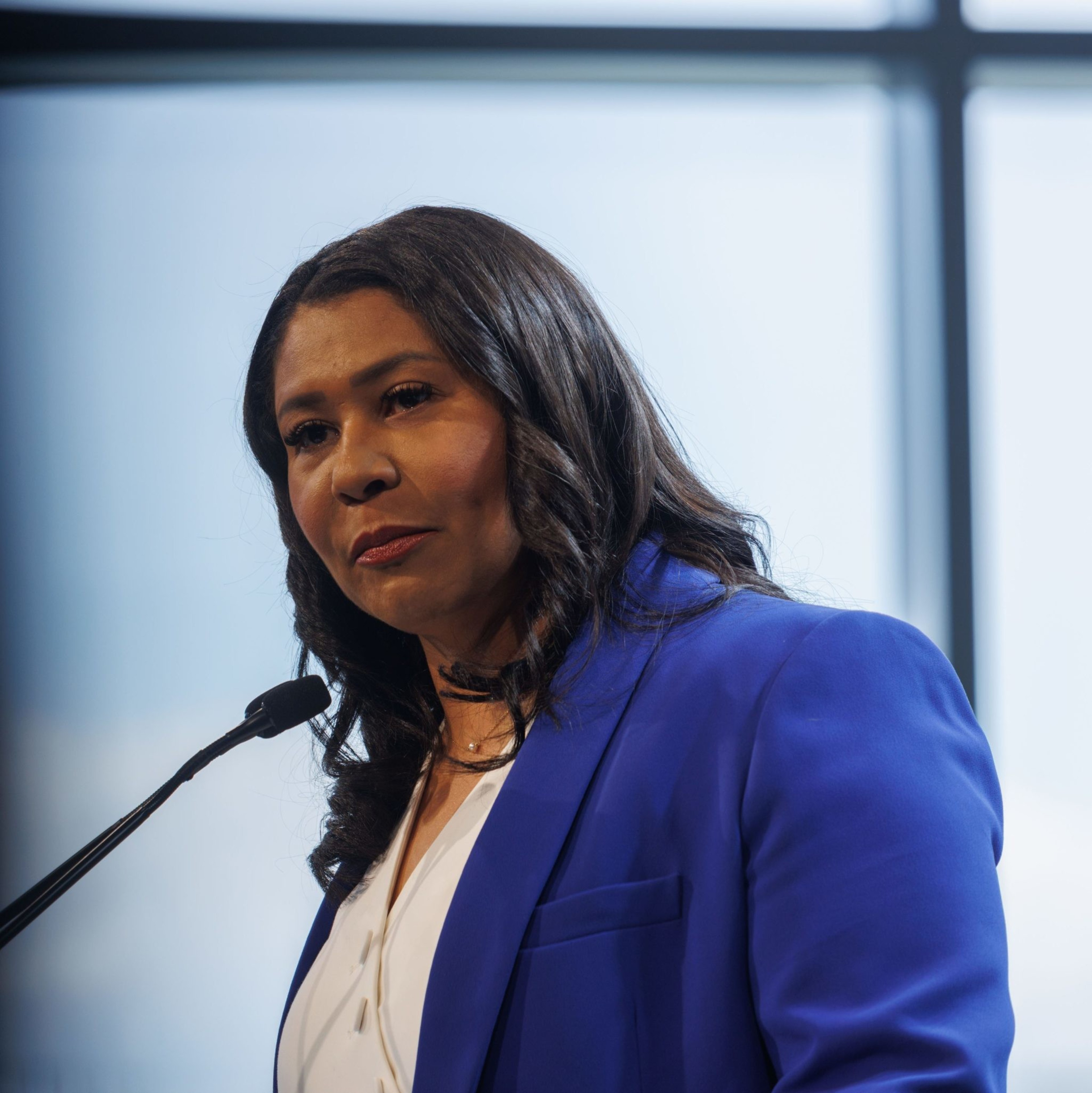 A woman in a blue blazer speaks at a podium, with large windows and soft light in the background.