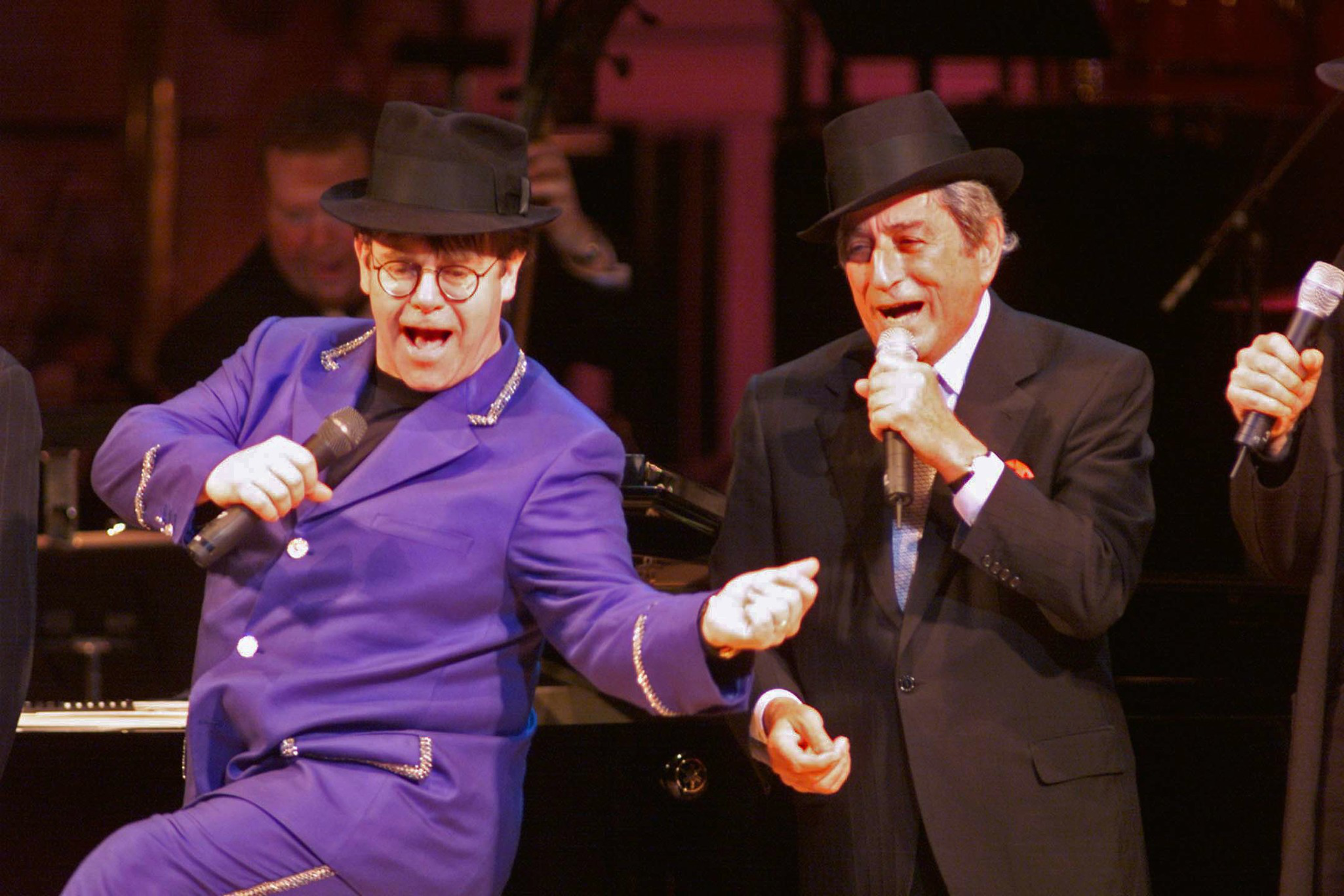 Two men wearing hats sing into microphones on stage, one in a bright purple suit and the other in a dark suit, both engaged in a lively performance.