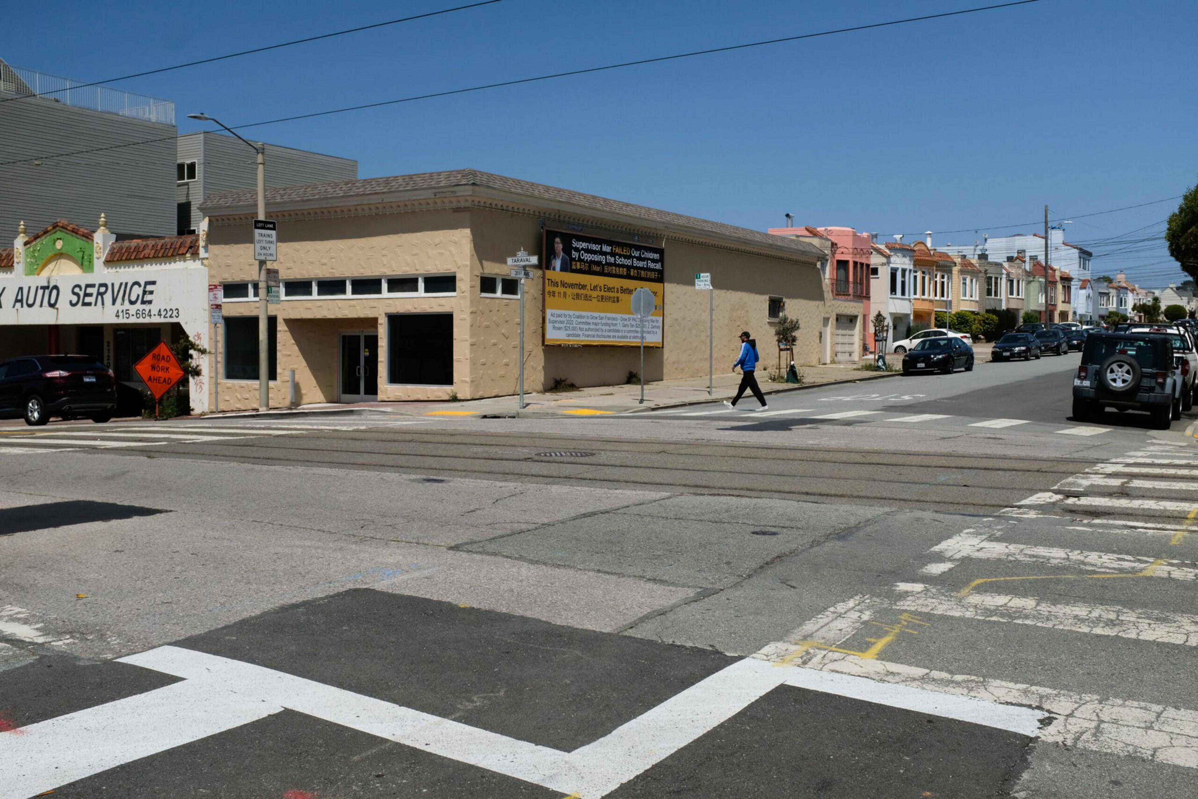 A person in blue walks at a quiet street corner with a beige building displaying a large sign, a car repair shop, and a row of colorful houses under a clear sky.
