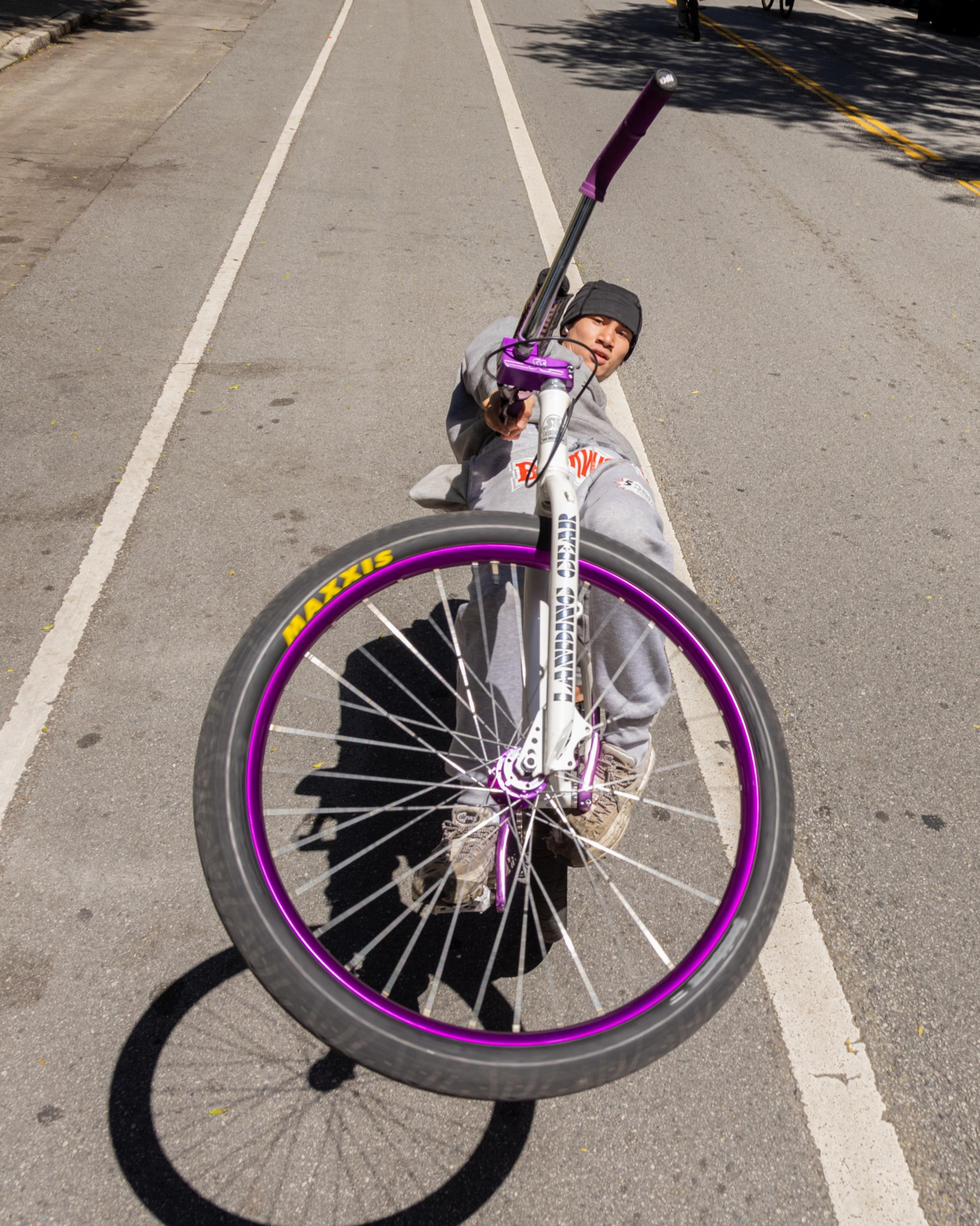 A person performs a wheelie on a bicycle with purple rims, wearing casual clothes and a beanie, on a sunny street with shadows cast on the ground.