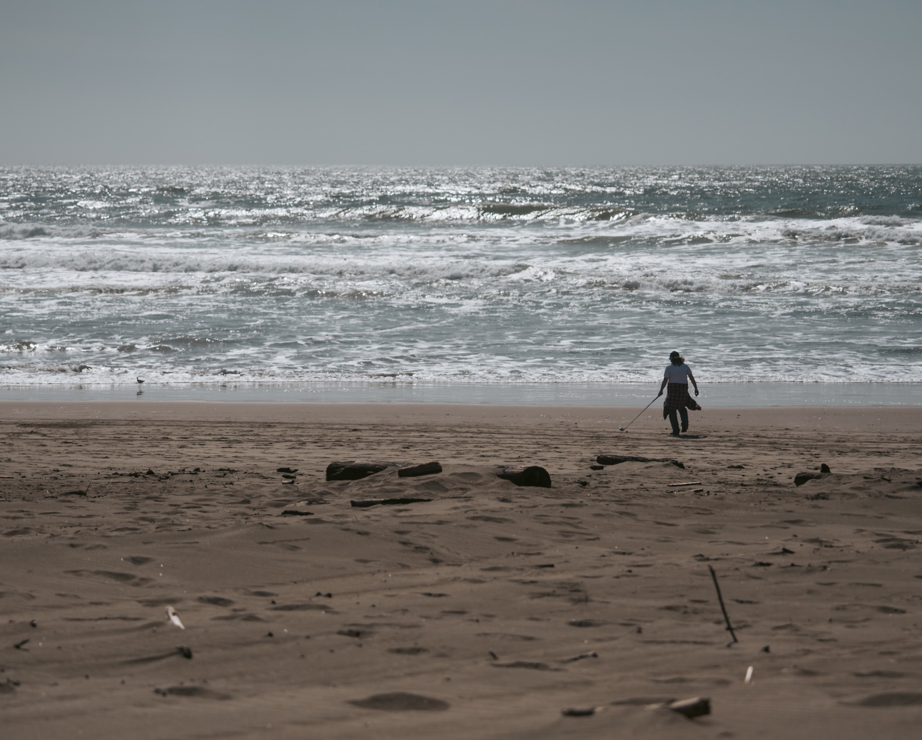A person walks alone on a sandy beach near the shore, holding a metal detector, with waves sparkling in the sunlight behind them.