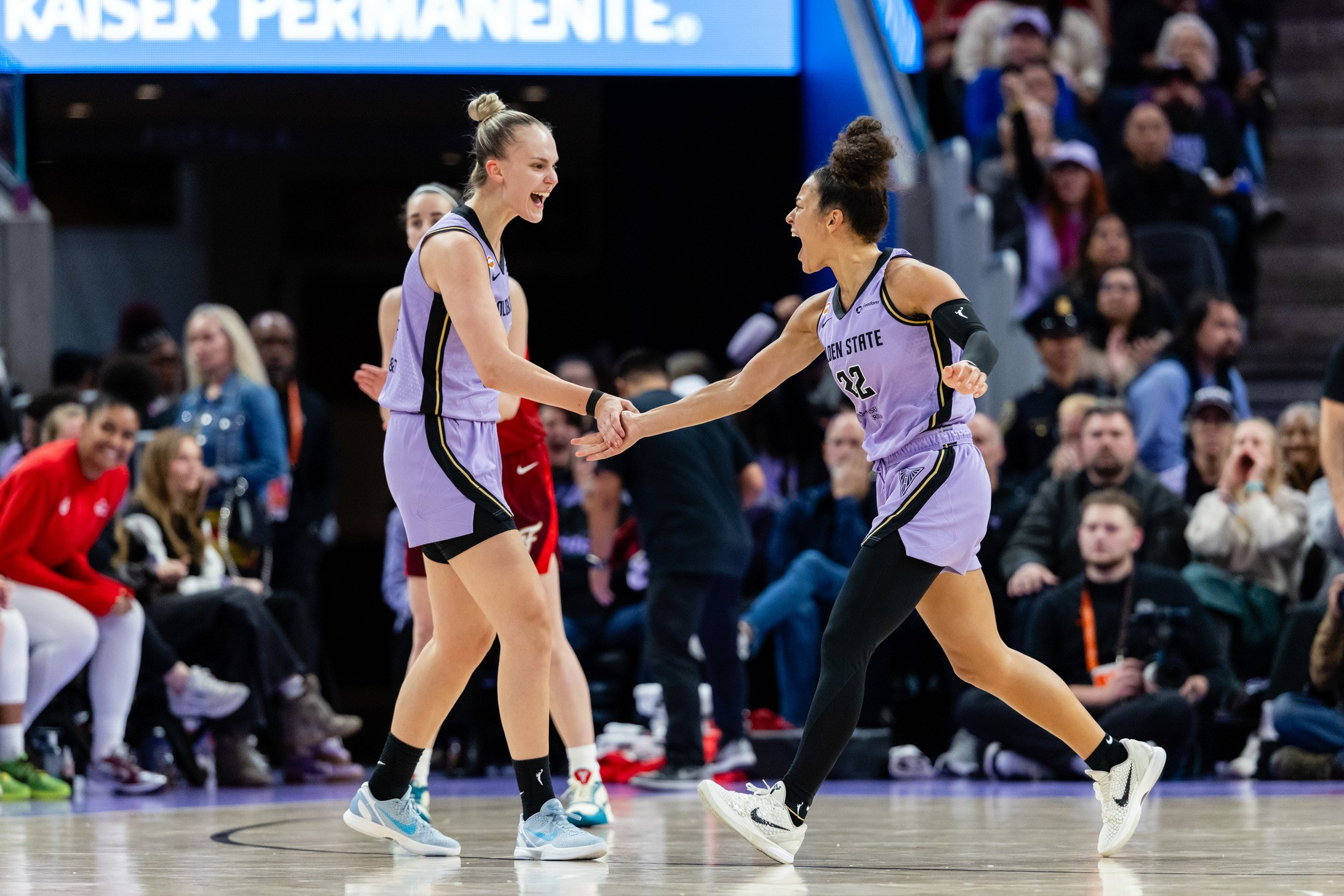 Two basketball players in purple Golden State uniforms celebrate with a handshake on the court while an audience watches intently.
