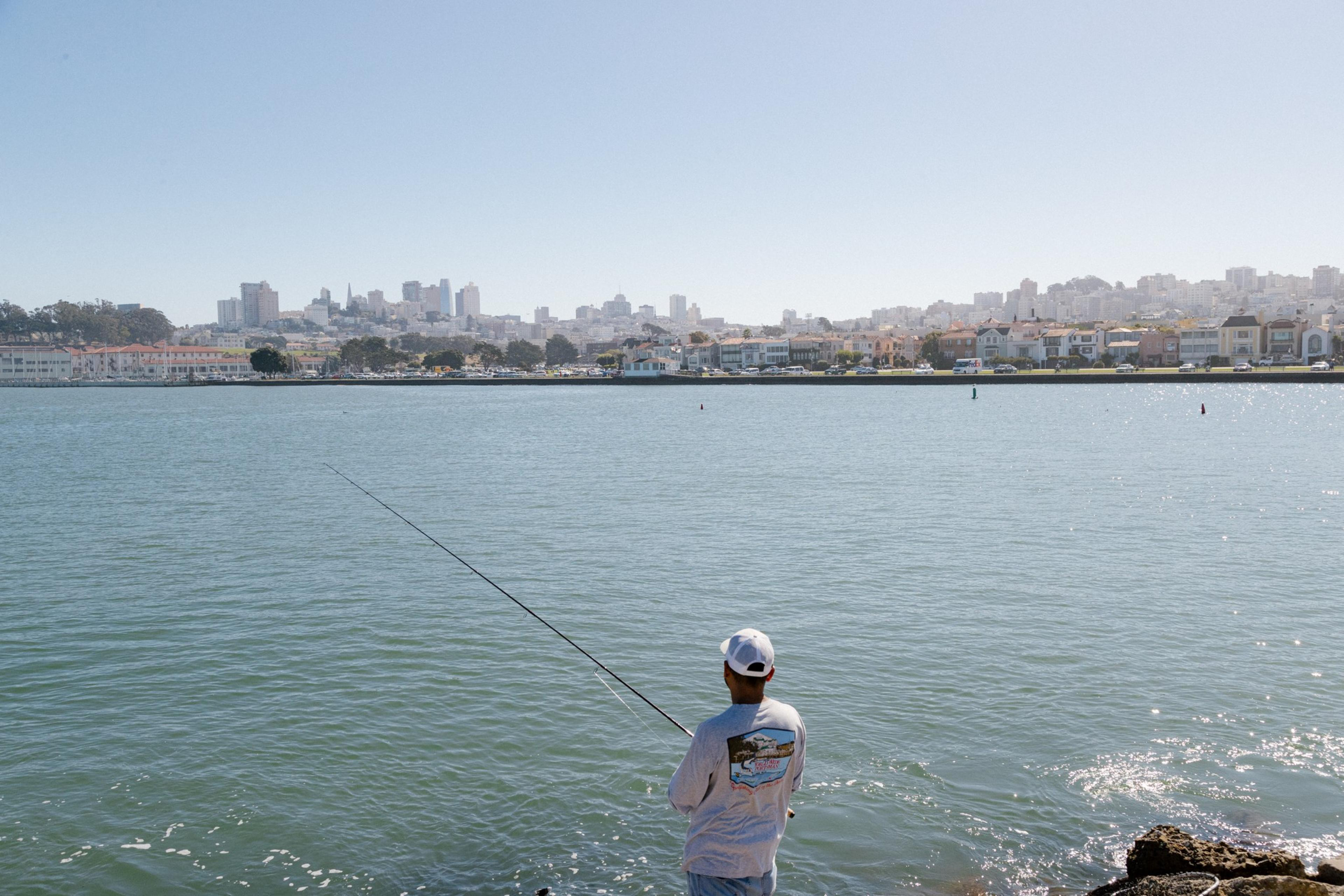 A person stands on rocky shore fishing in calm water with a city skyline and residential buildings visible across the bay under a clear sky.