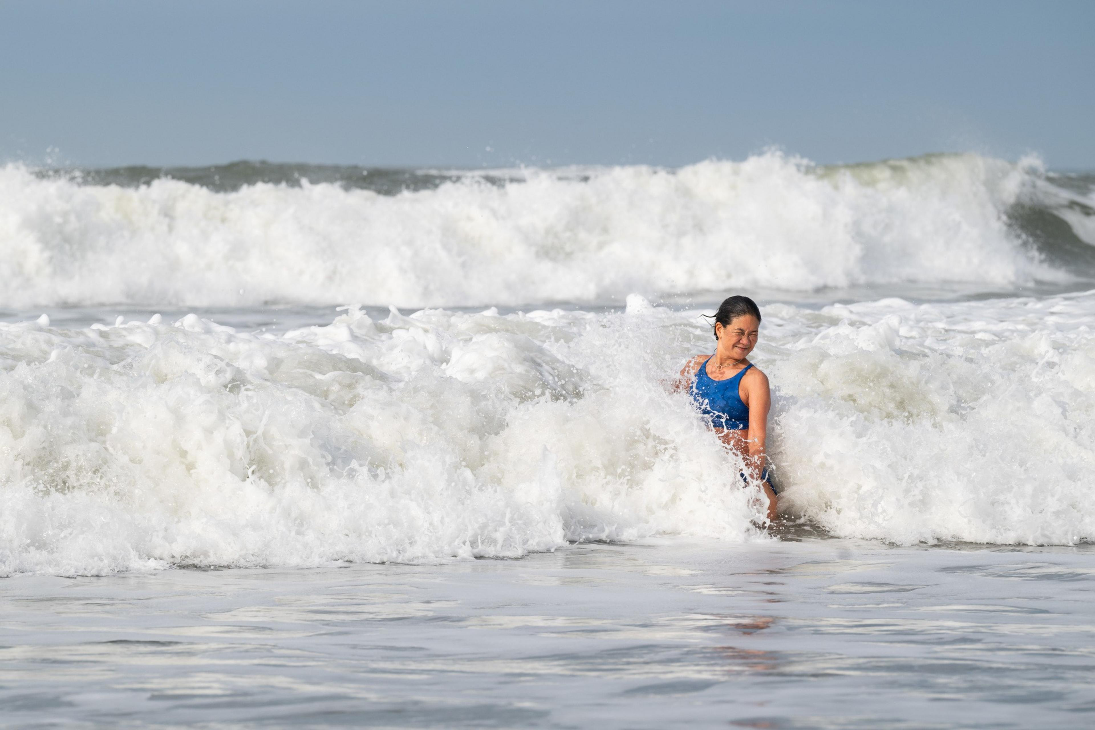 A woman in a blue swimsuit is joyfully standing in foamy ocean waves, with more waves rolling in. The sky above is clear and blue.