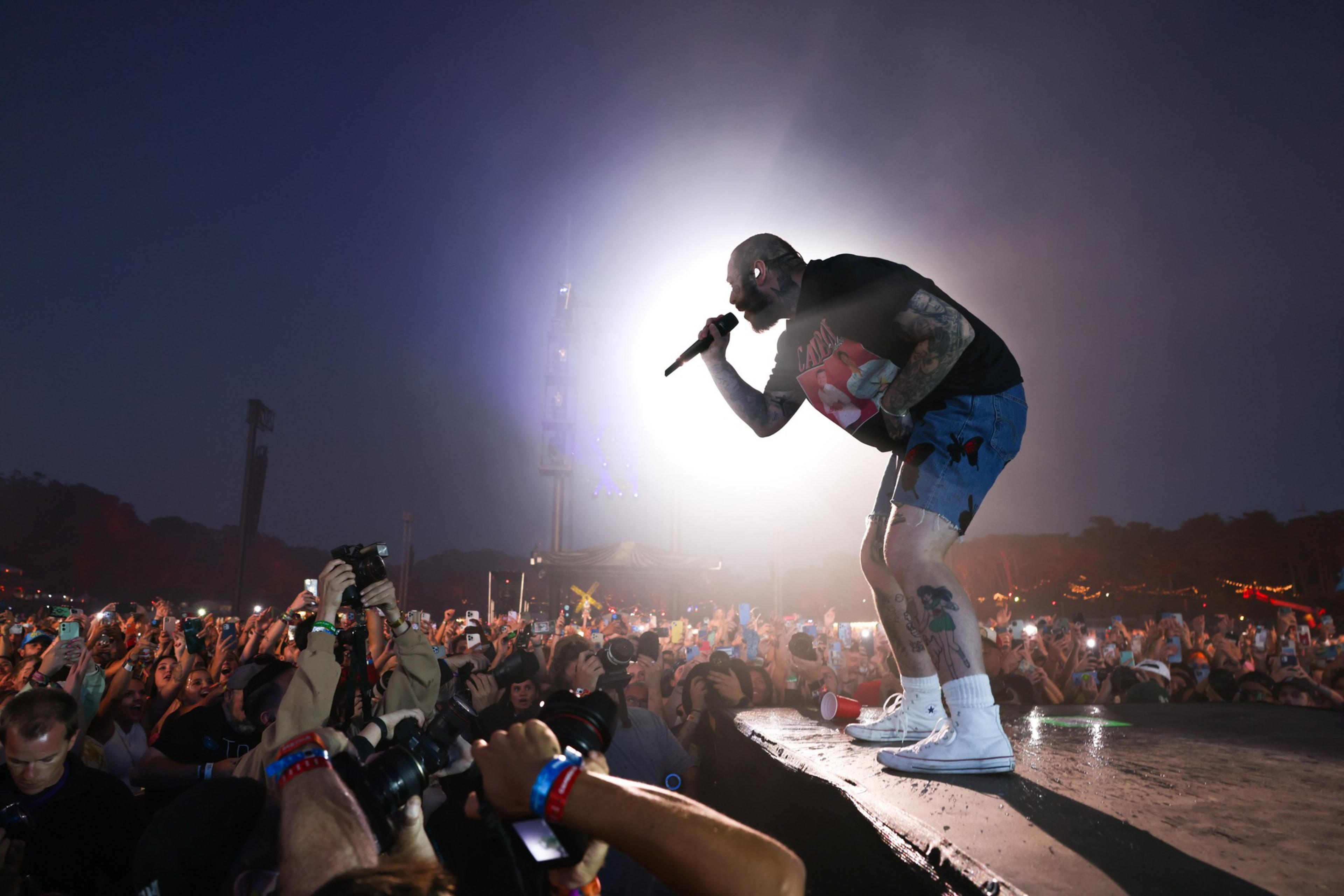 A tattooed singer in a black t-shirt and blue shorts performs on stage at night, leaning forward with a microphone toward an enthusiastic crowd.
