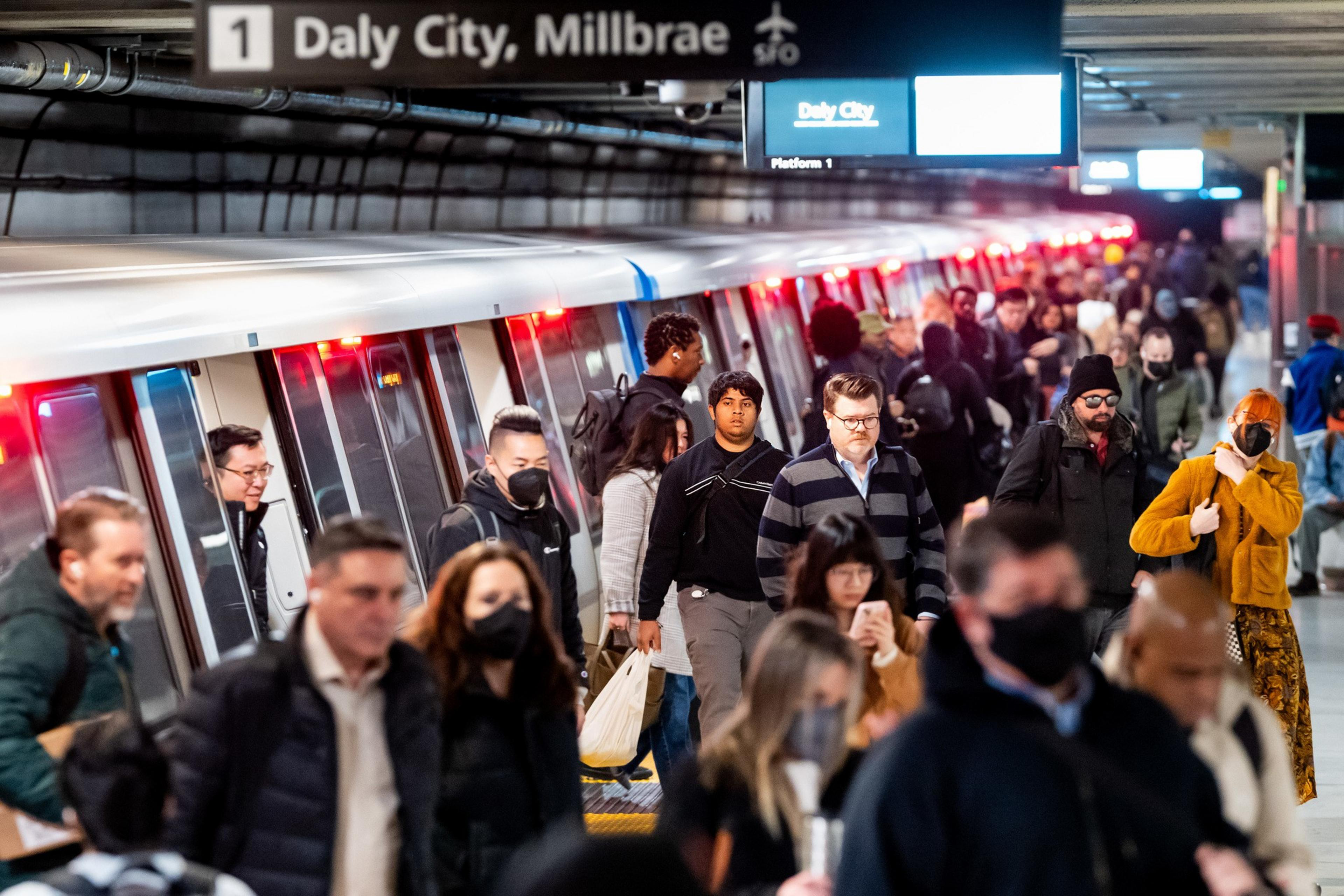 The image shows a crowded subway platform with a train labeled &quot;Daly City, Millbrae.&quot; Commuters, some wearing masks, are walking in various directions.