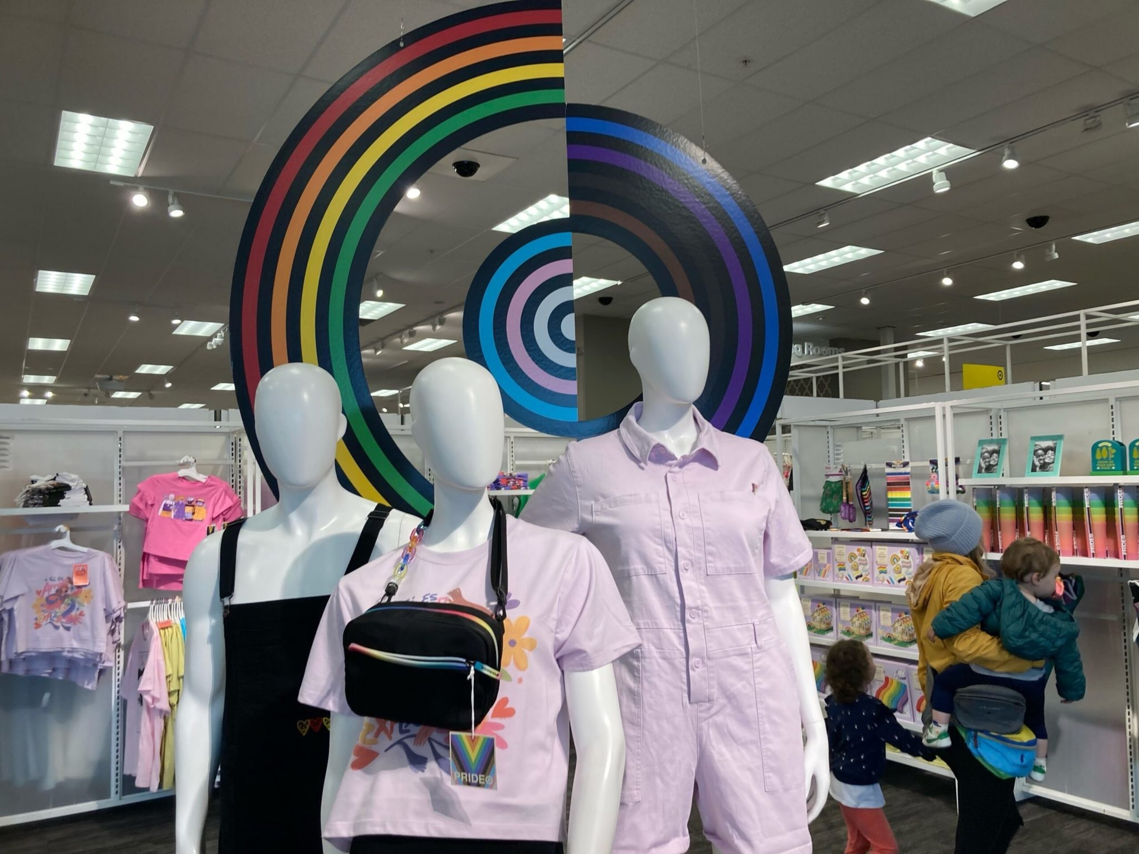 Three faceless mannequins wear pastel clothing and rainbow accessories beneath a large, colorful circular art piece in a brightly lit store.