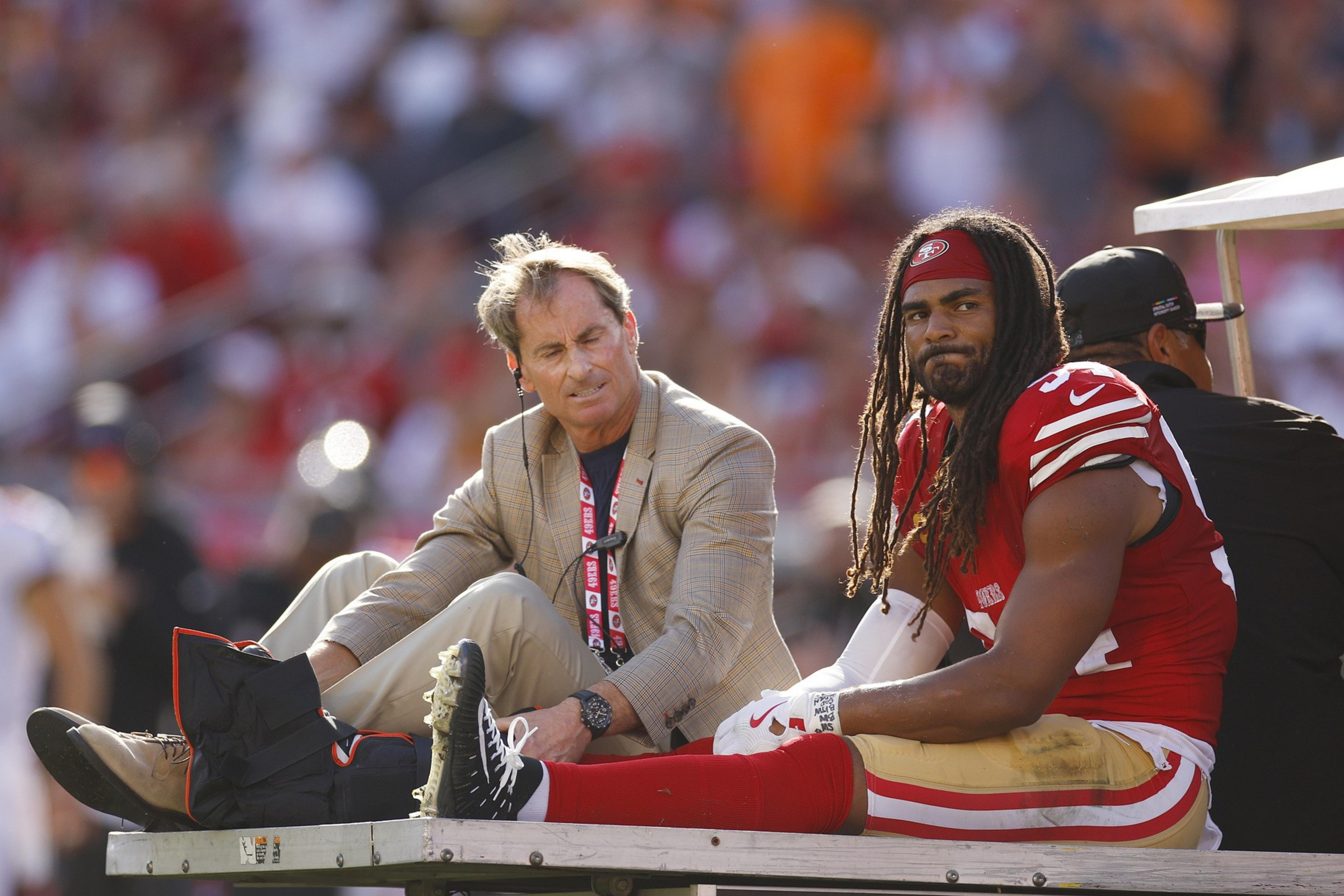 A man in a beige suit with a black boot on his left foot sits next to a football player wearing a red and gold uniform and a red headband, both on a cart.
