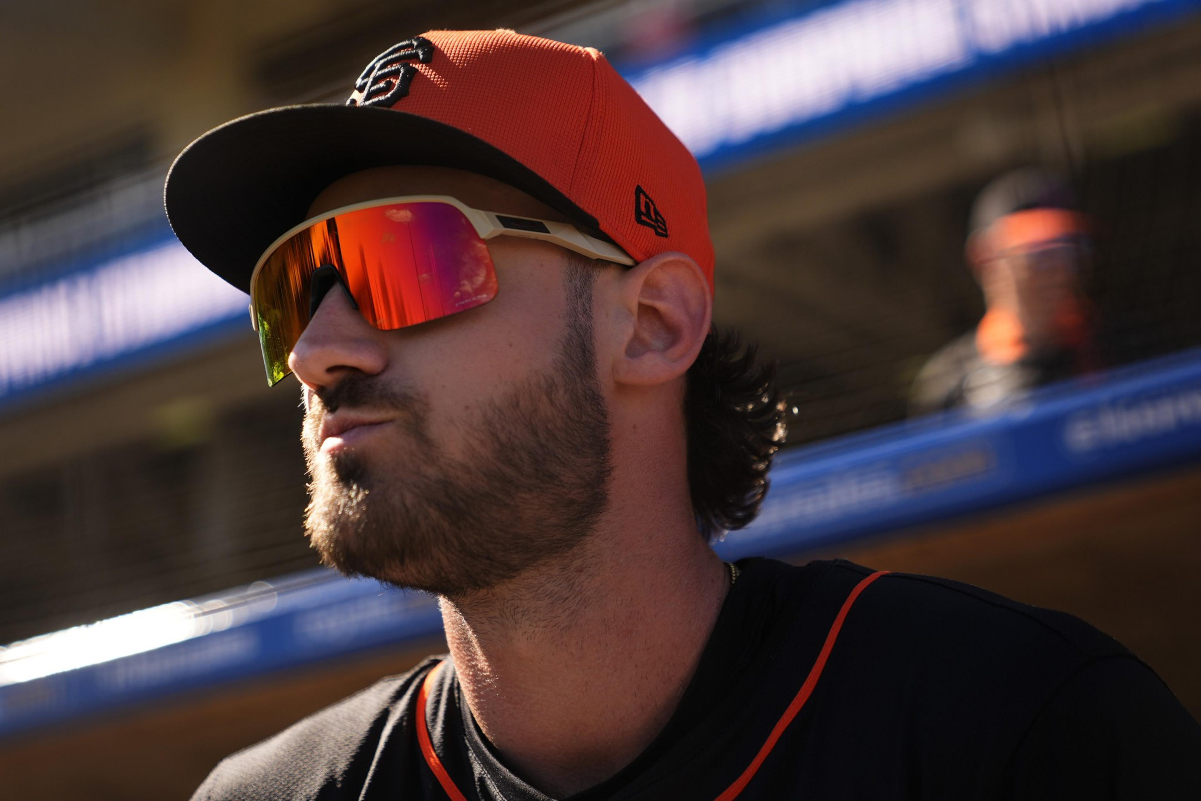 A person wearing an orange baseball cap and reflective sunglasses looks to the side. They have facial hair and wear a black sports shirt with orange trim.