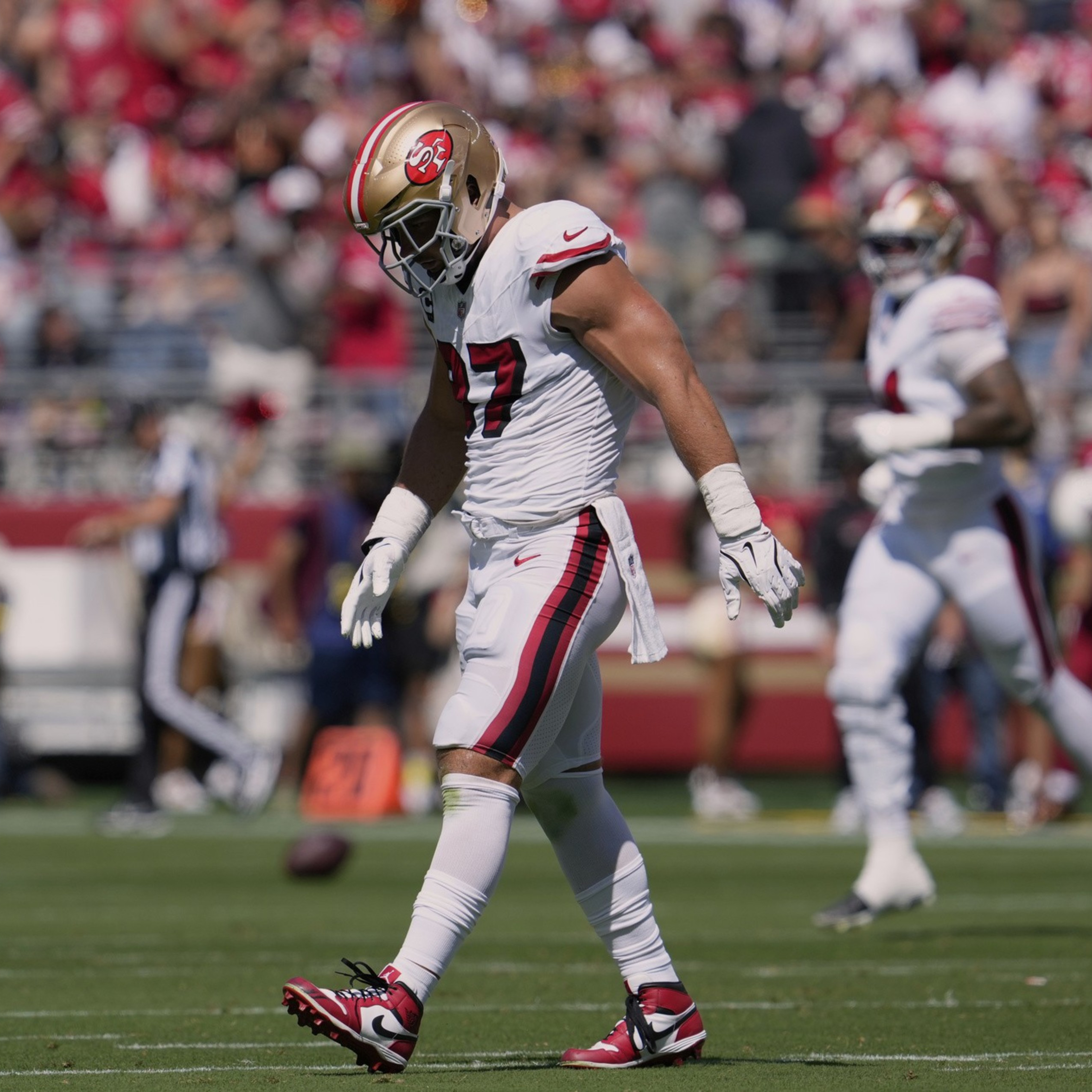 A football player wearing a white and red San Francisco 49ers uniform walks on the field with his head down during a game.