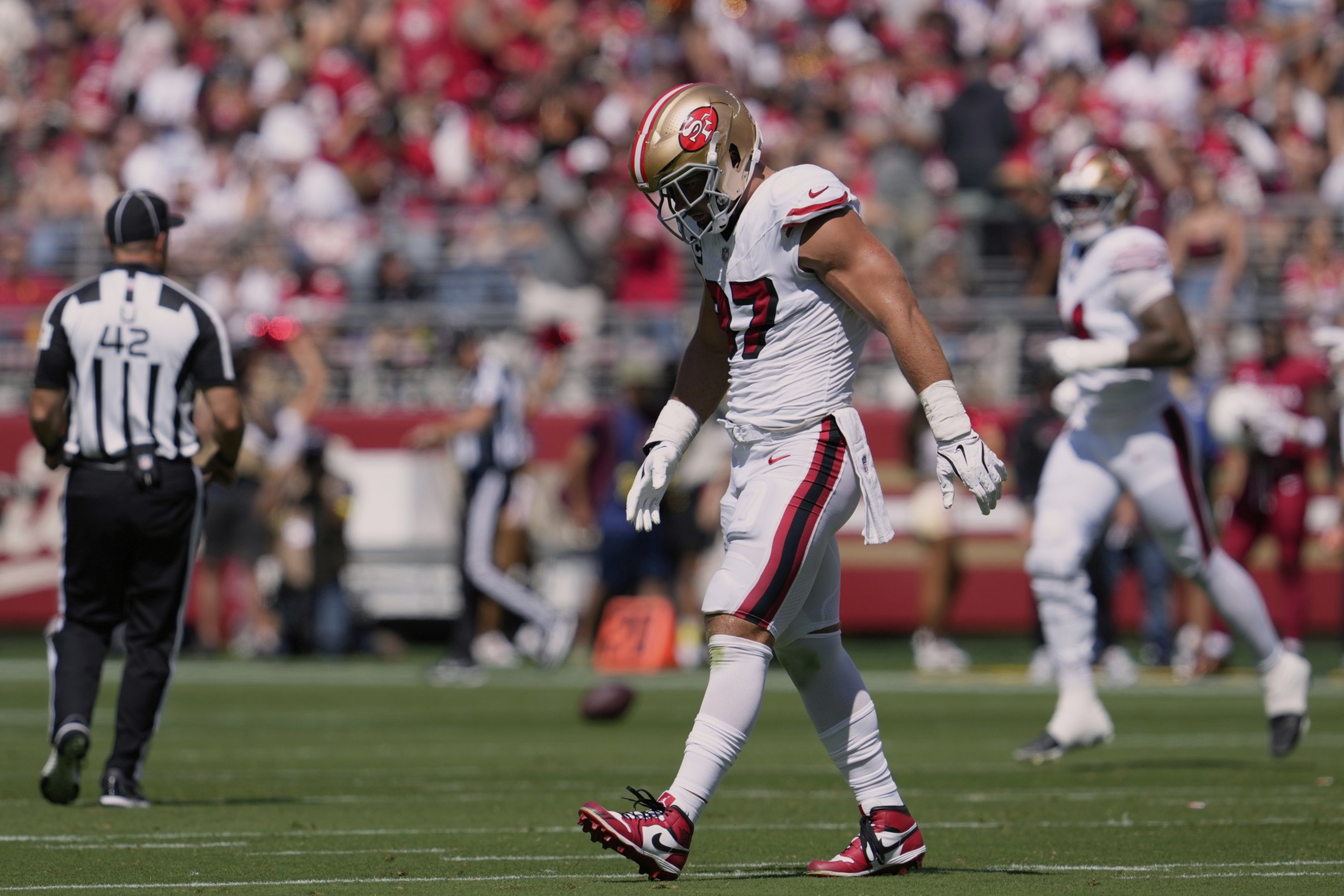 A football player wearing a white and red San Francisco 49ers uniform walks on the field with his head down during a game.