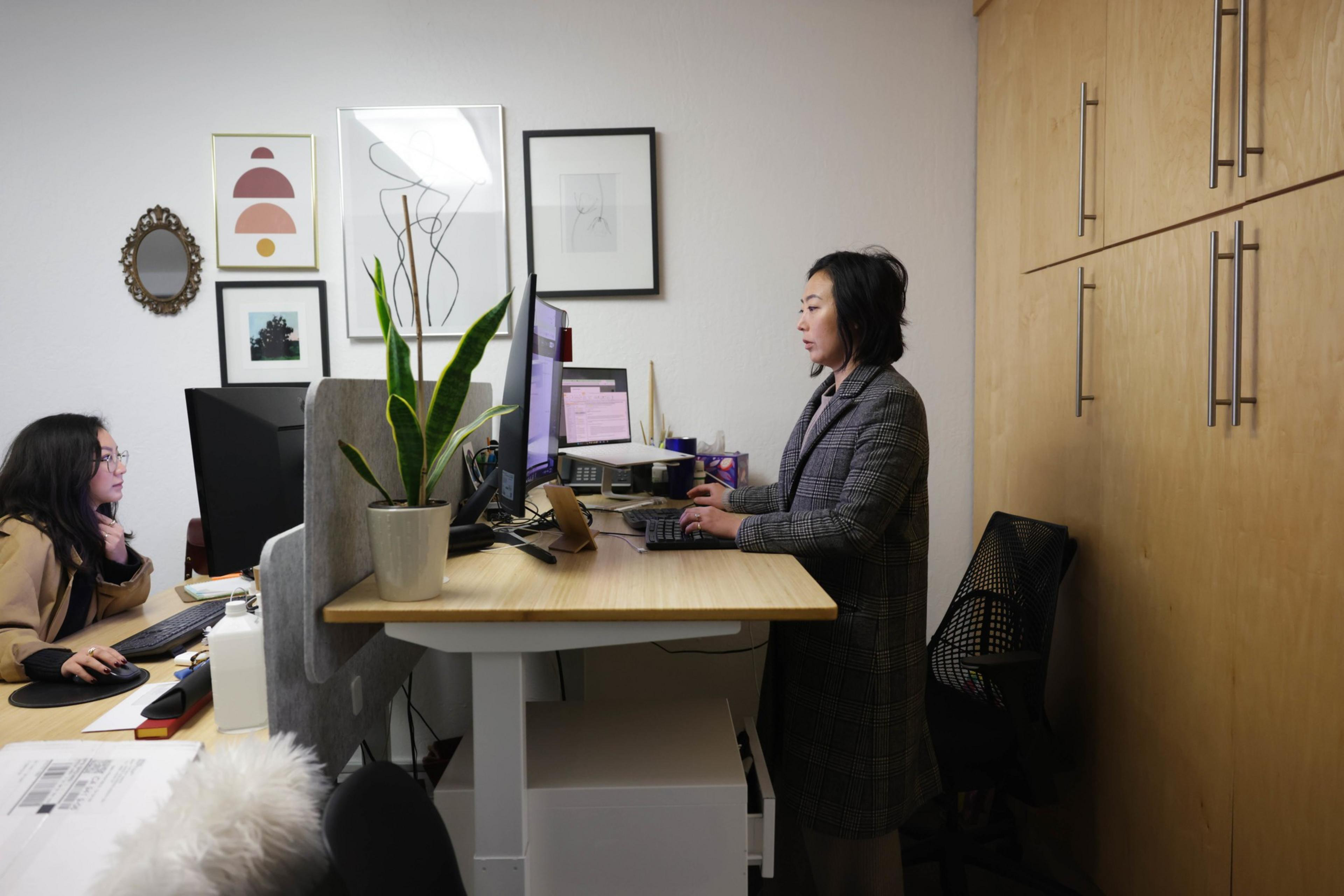 A person at their desk in a gray coat jacket types at a computer.