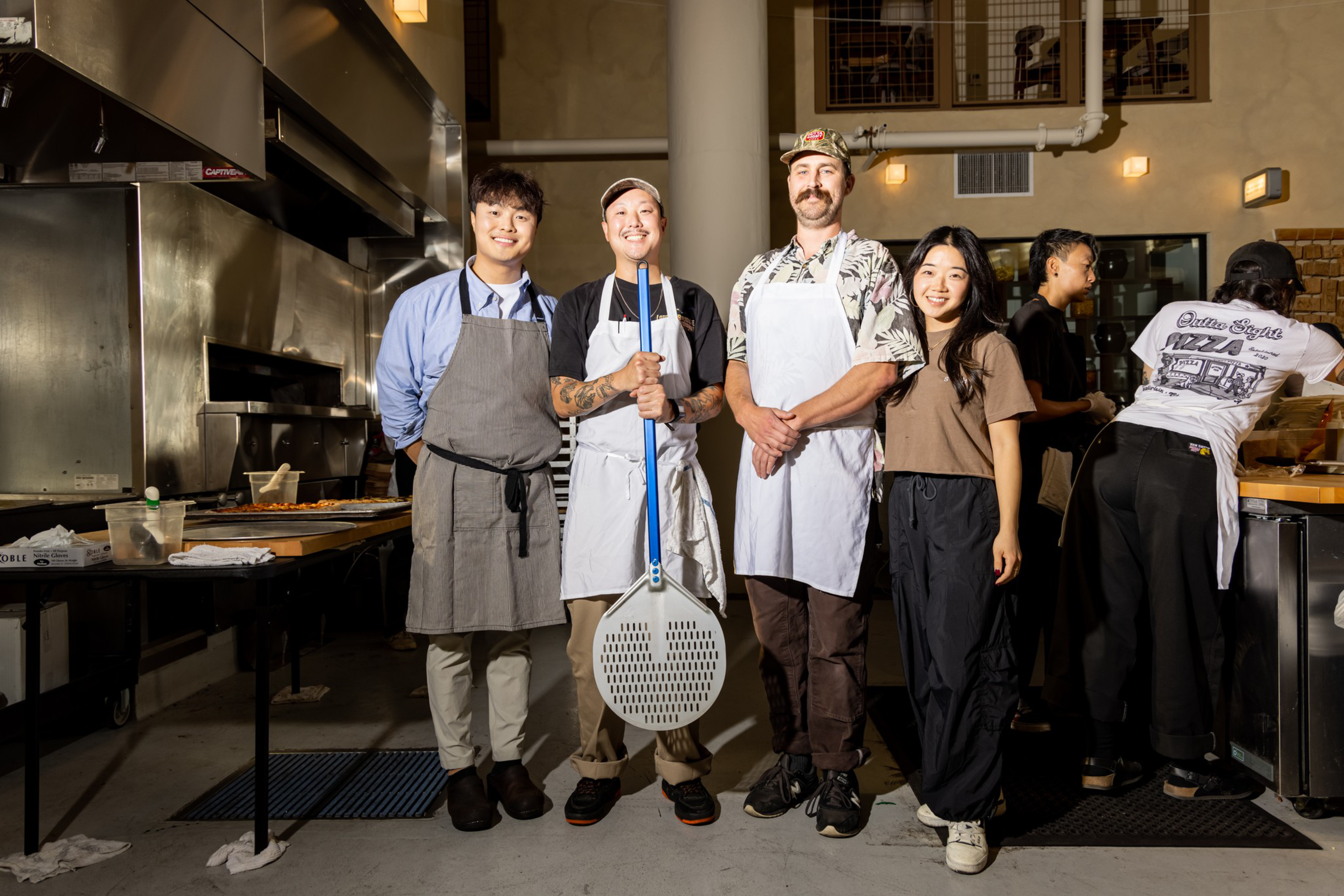 Four smiling people stand in a kitchen, three wearing aprons and one holding a pizza peel, with two others working in the background.
