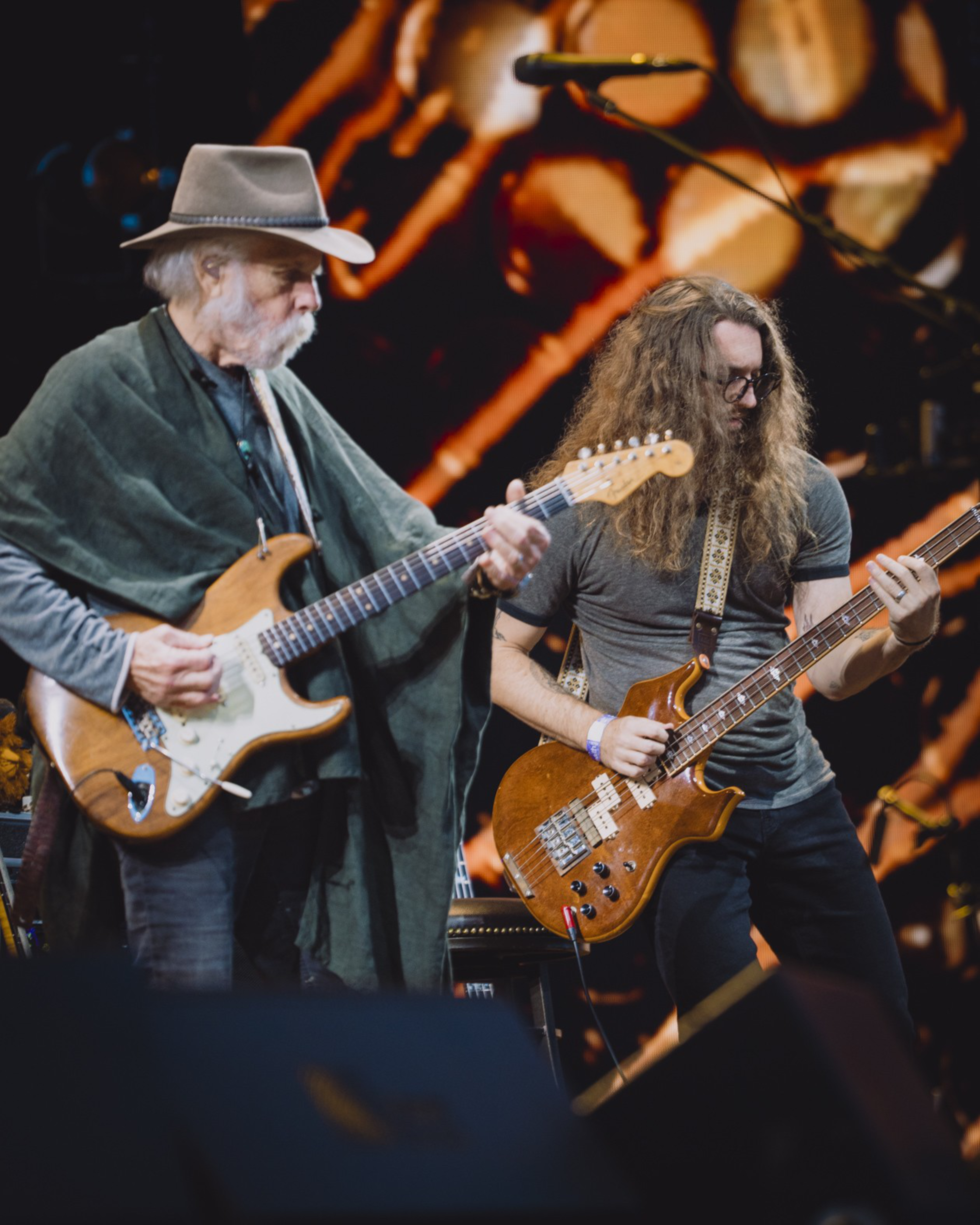 An older man with a gray beard and hat plays electric guitar beside a long-haired man in glasses playing bass on stage with a warm, blurry background.