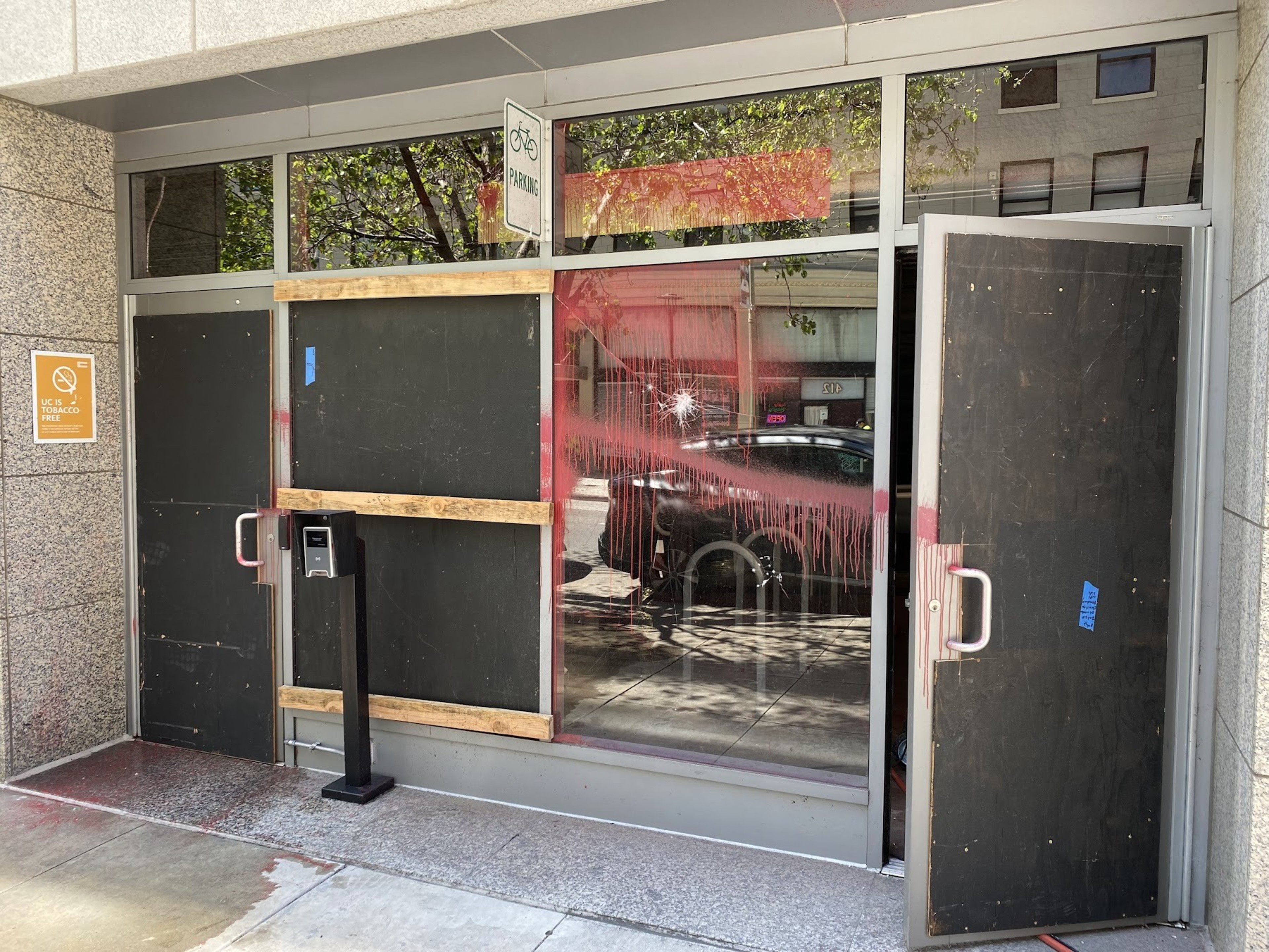 The image shows a storefront with boarded-up doors, reflecting a bright day, situated under a grey stone archway. Signs warn of no parking and indicate a tobacco-free zone.