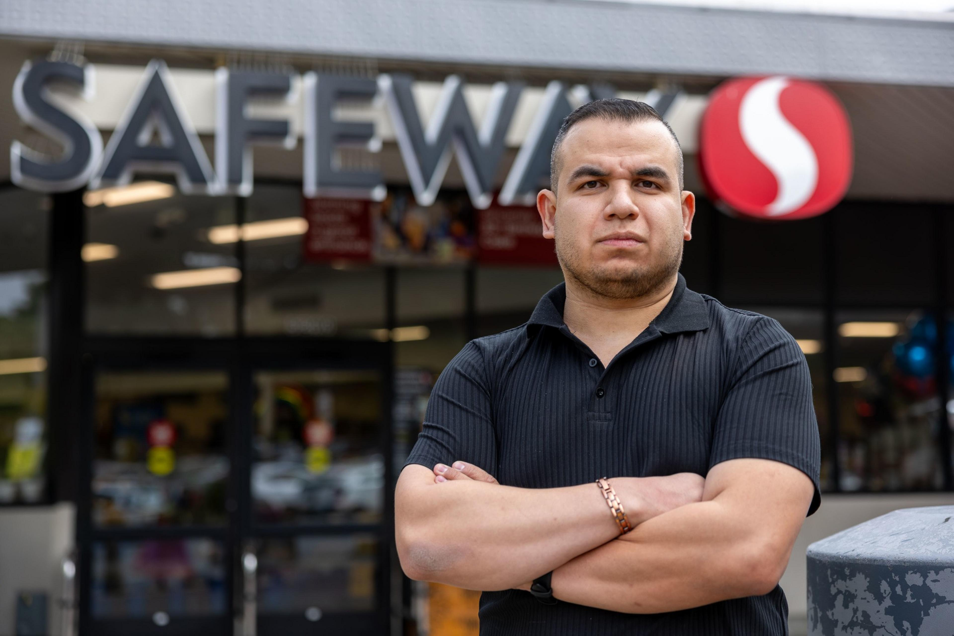 A man stands with arms crossed in front of a Safeway supermarket. He is wearing a black polo shirt and appears serious, with the store's signage clearly visible above him.